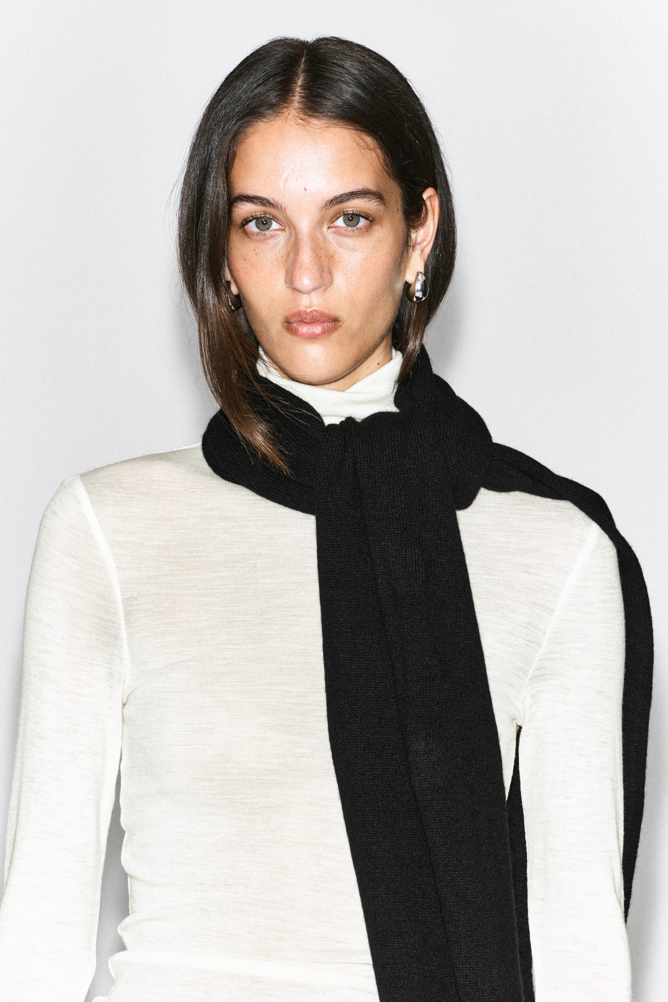 A black cashmere scarf is being worn by the model, wrapped once around the neck over a cream-coloured turtleneck top. One end of the fine-knit scarf hangs straight down the front, while the other end drapes over the shoulder.