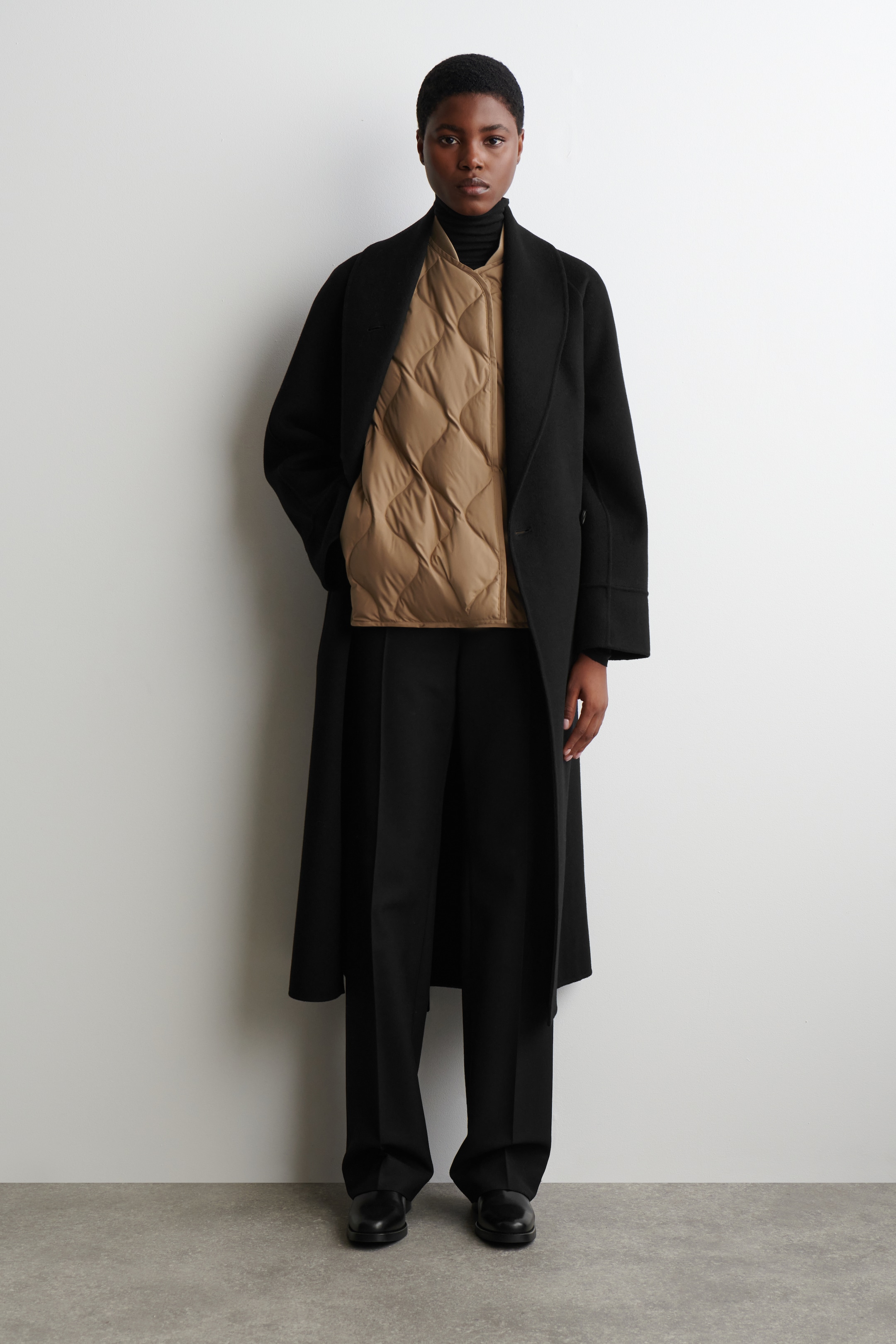 View larger image: A long black coat is worn open, featuring broad lapels and a relaxed silhouette, revealing a tan quilted vest with large diamond-shaped quilting. The vest is layered over a black turtleneck and wide-leg black pants, which are worn with black shoes.