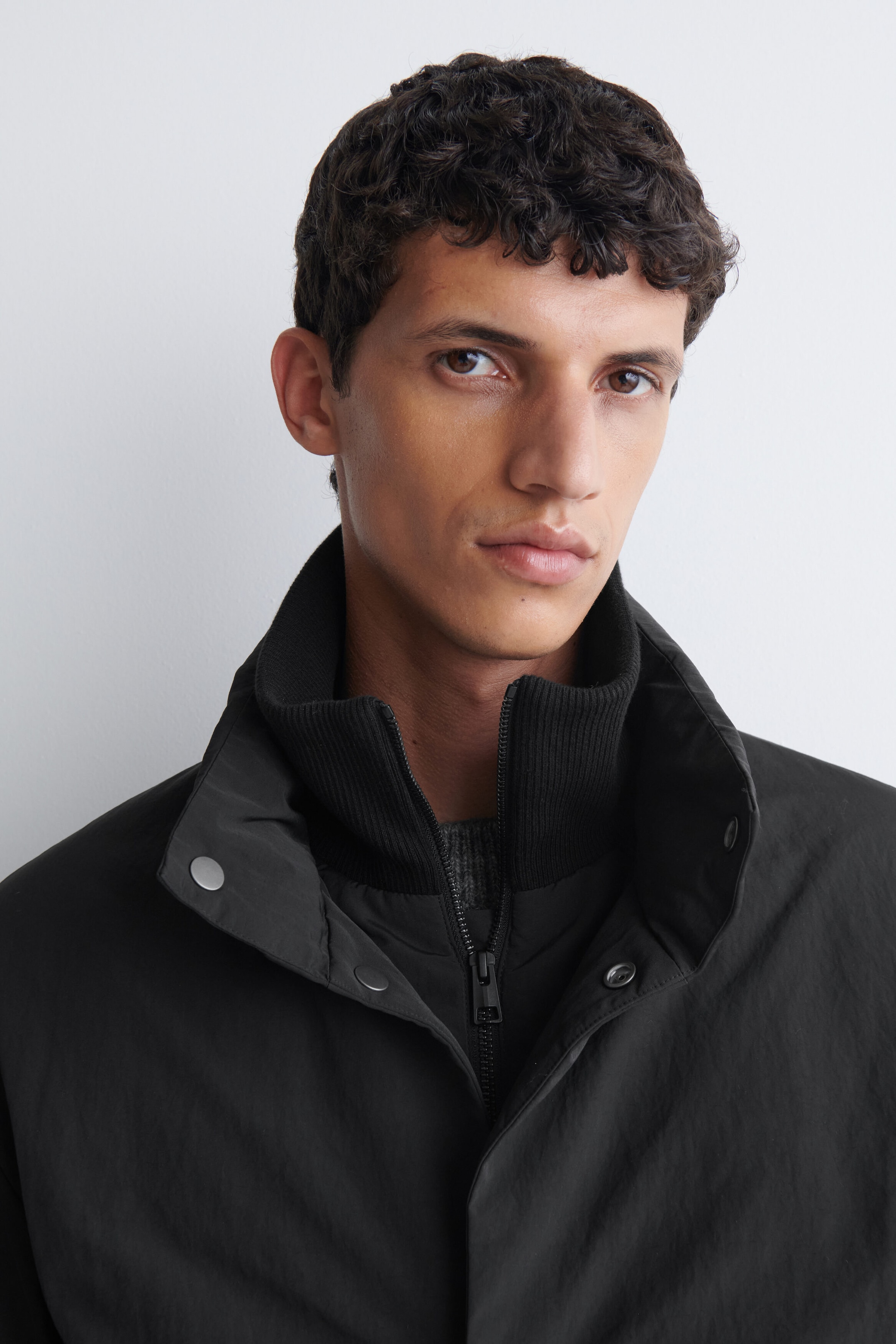 The model is wearing a black padded funnel-neck down jacket, featuring a double-collar construction with a zippered, ribbed inner collar and a snap-button outer collar. The jacket is layered over a grey knit garment. The model is wearing a black padded funnel-neck down jacket, featuring a double-collar construction with a zippered, ribbed inner collar and a snap-button outer collar. The jacket is layered over a grey knit garment.