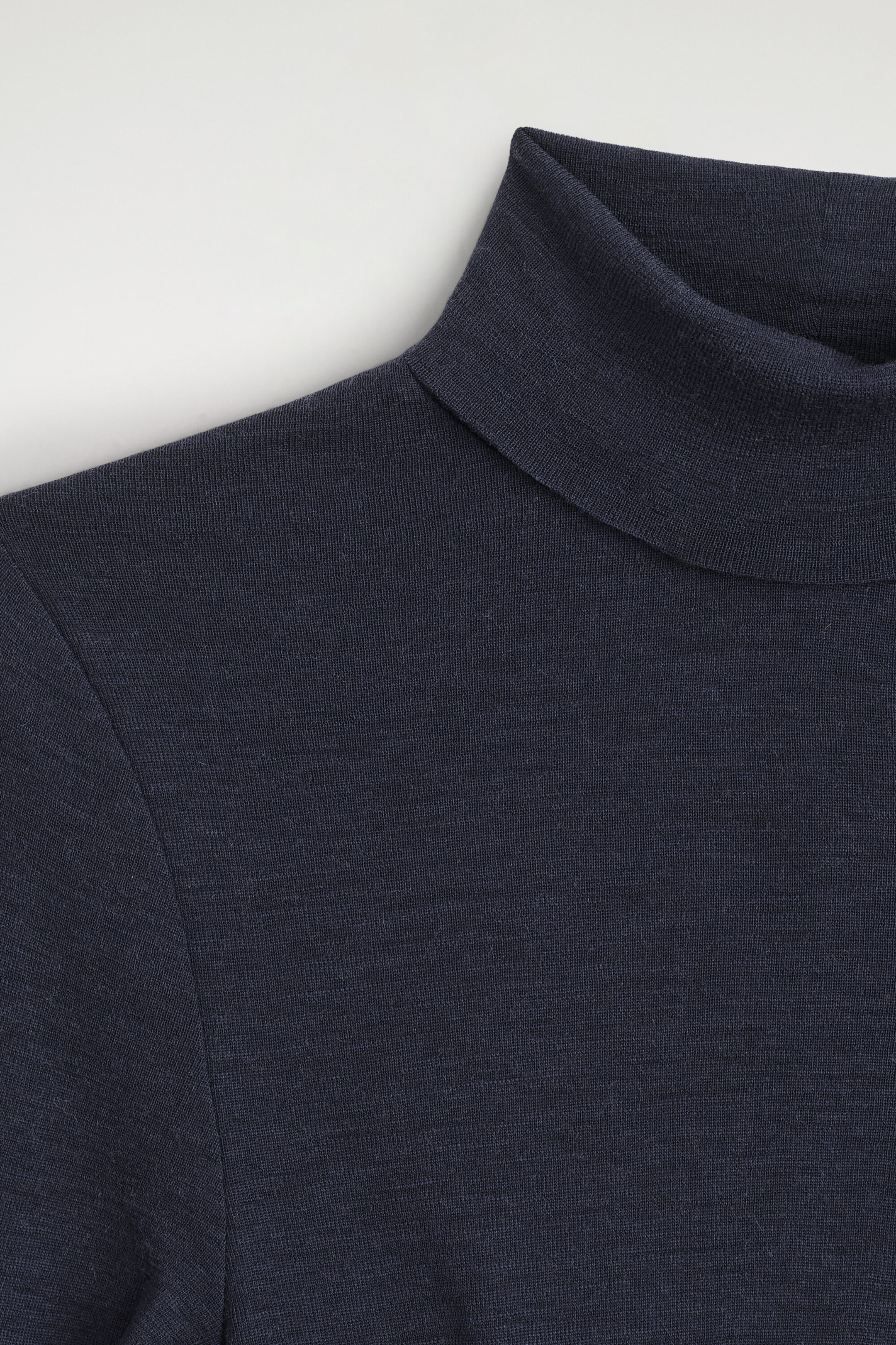The merino wool high-neck top is shown in a close-up front view, highlighting its fine-knit texture and folded mock neck collar in a dark navy blue color.