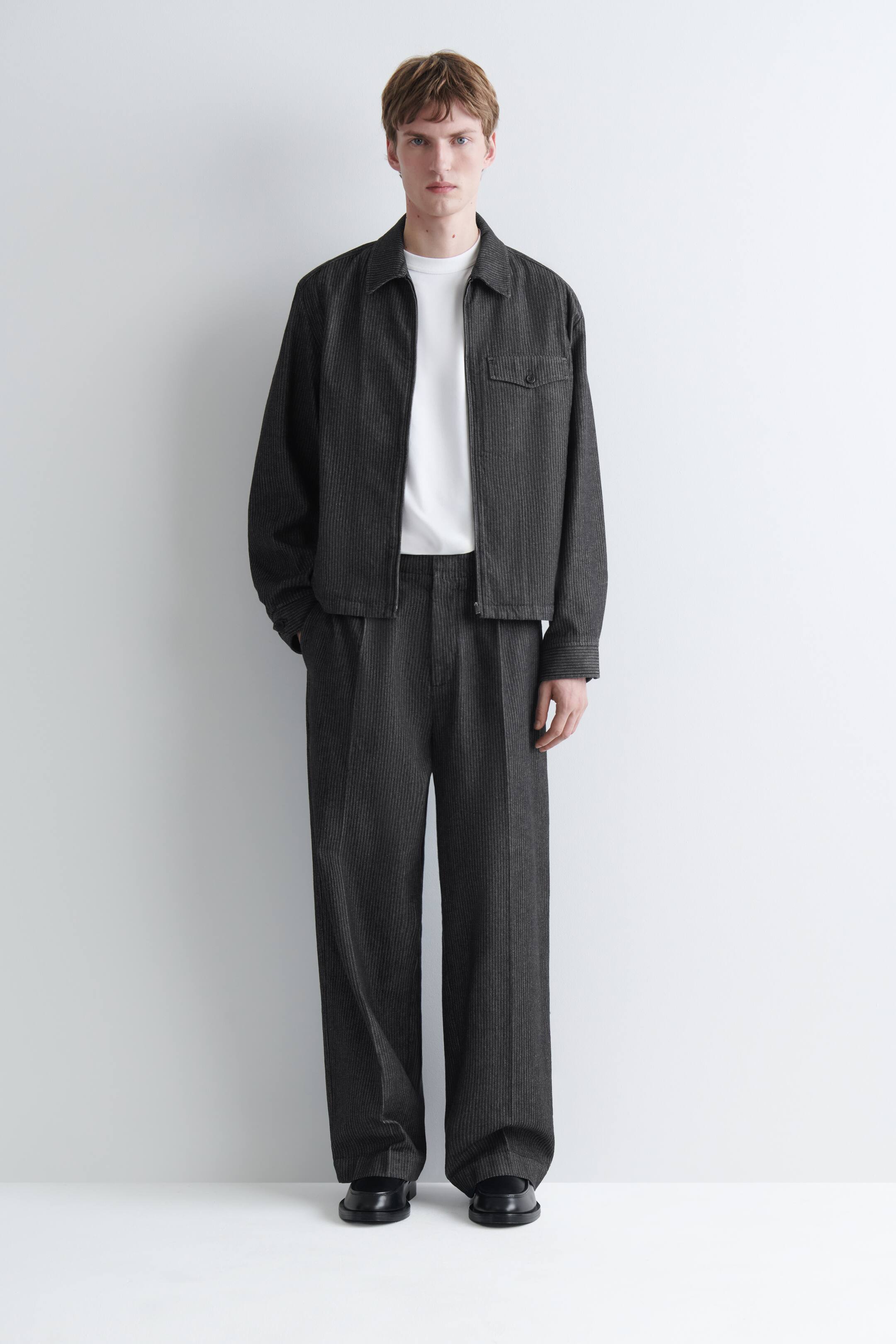Agrandir l'image: Ce pantalon en denim à fines rayures, coupe ample et jambe large, de couleur gris foncé, est porté par le mannequin, qui a une main posée dans la poche. Le pantalon présente des plis sur le devant et une silhouette décontractée, et il est associé à une veste zippée assortie, à un t-shirt blanc rentré à l’intérieur et à des chaussures noires.