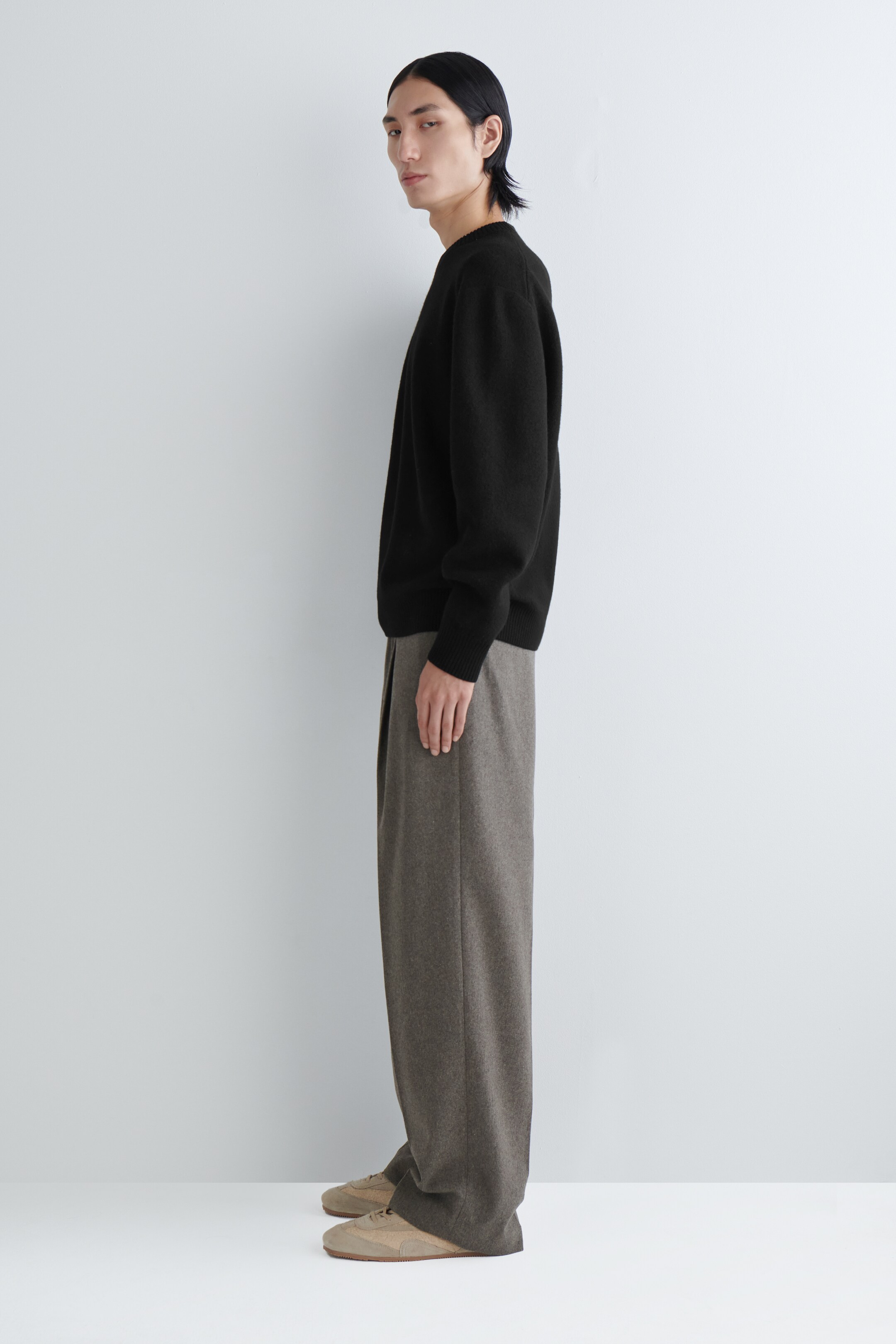 Grotere afbeelding bekijken: Een zwarte gebreide trui met lange mouwen wordt in zijaanzicht gepresenteerd, met geribde boorden en een ronde hals. Het wordt gestyled met gemêleerde bruine wijde broek, die een ontspannen pasvorm heeft met een zichtbare voorplooi, en lichtbruine lage sneakers.