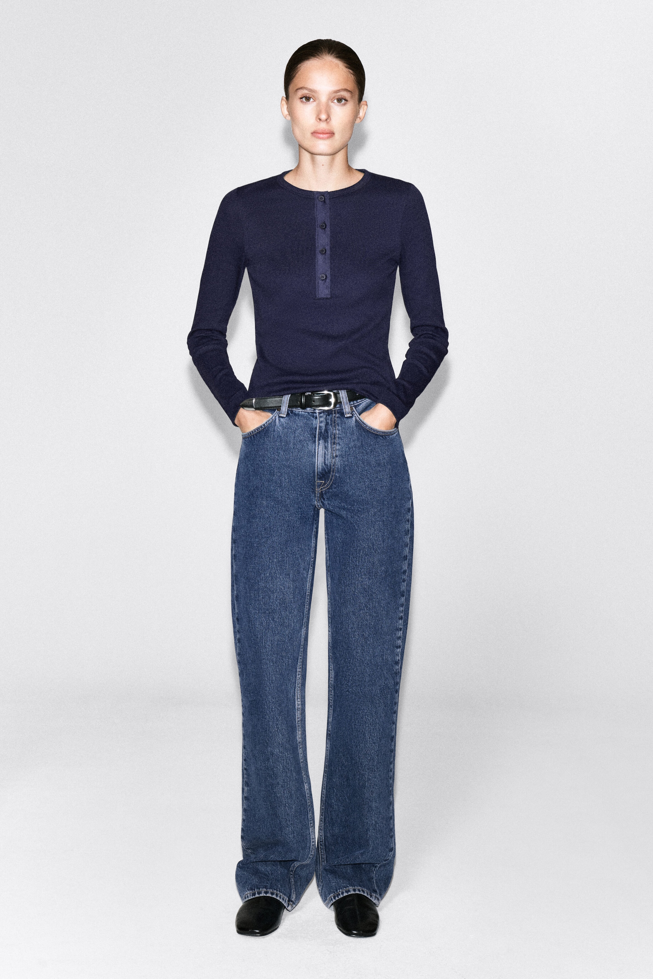 A navy blue ribbed cotton Henley top with a buttoned placket and long sleeves is worn by the model. The top is styled tucked into classic blue jeans with a black belt. A navy blue ribbed cotton Henley top with a buttoned placket and long sleeves is worn by the model. The top is styled tucked into classic blue jeans with a black belt.