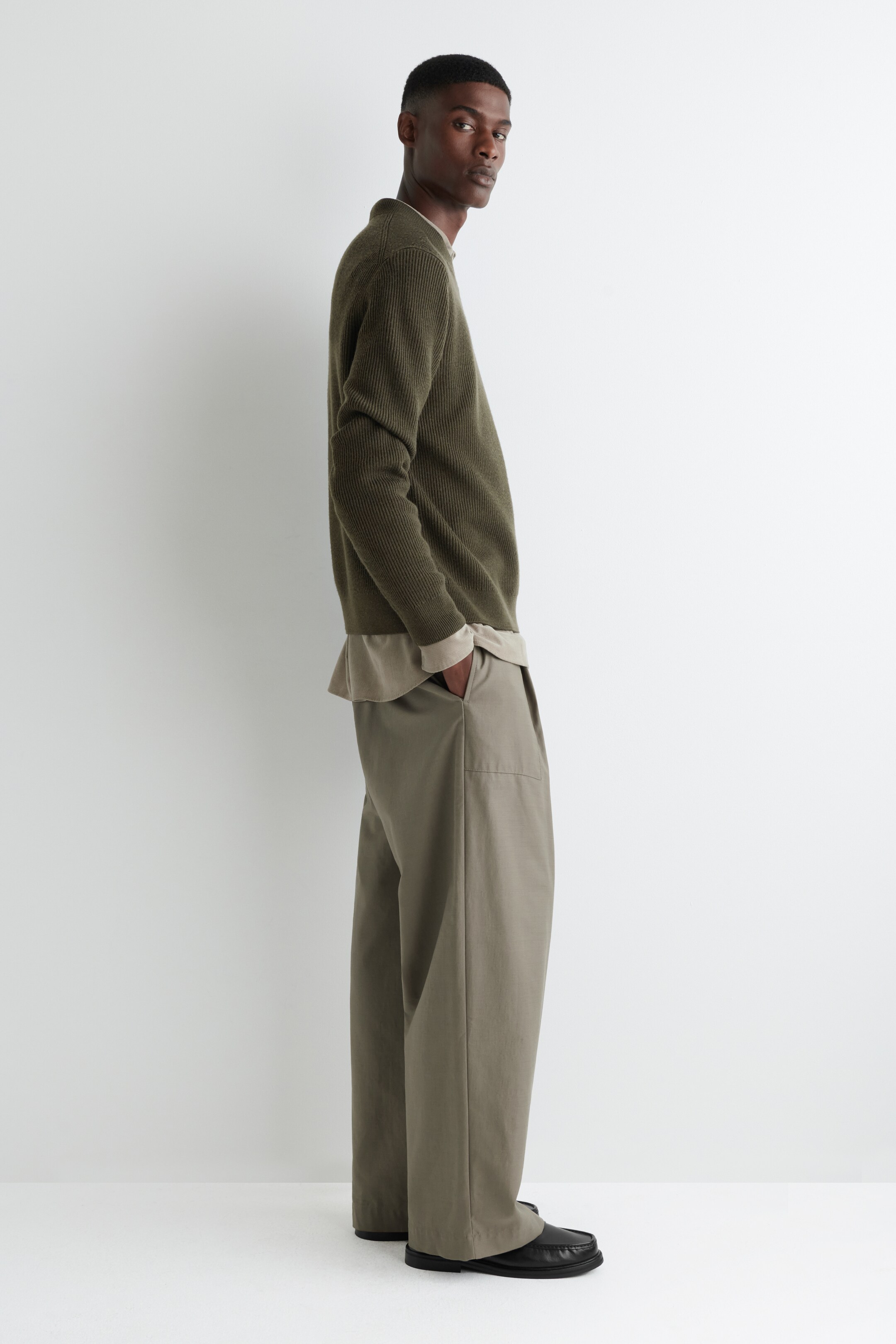 Visualizza immagine più grande: Un maglione a coste in maglia verde oliva, con scollo rotondo e polsini a coste, è stratificato su una camicia beige chiaro, con entrambi i capi che mostrano una vestibilità rilassata in un profilo laterale. Il maglione è abbinato a pantaloni khaki a gamba larga, che presentano tasche laterali visibili, e scarpe nere senza lacci completano l'ensemble.
