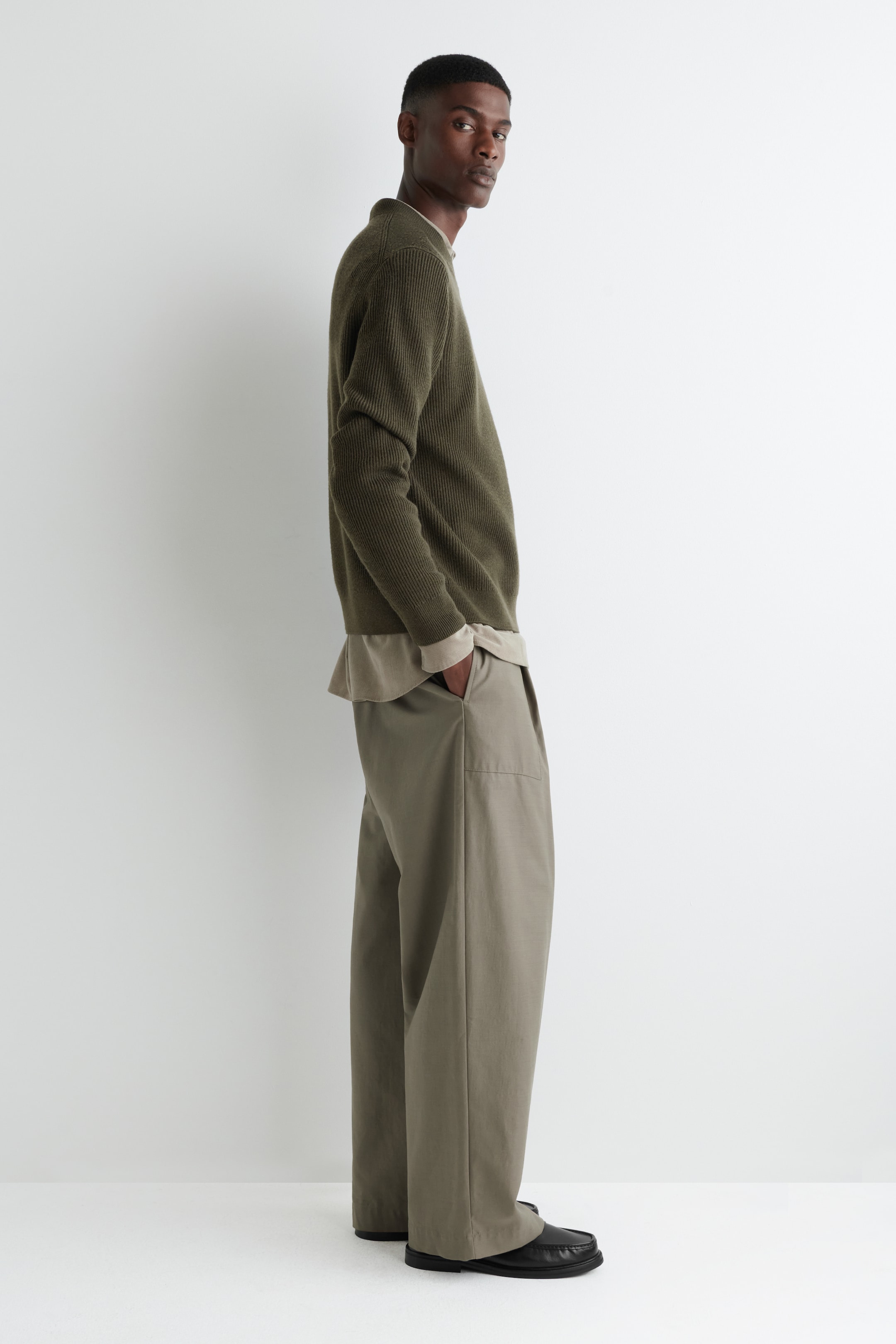 Ver imagen más grande: Un suéter de punto acanalado verde oliva, con cuello redondo y puños acanalados, está superpuesto sobre una camisa beige claro, con ambas prendas mostrando un ajuste relajado en un perfil lateral. El suéter se combina con pantalones caqui de pierna ancha, que presentan bolsillos laterales visibles, y unos zapatos negros sin cordones completan el conjunto.