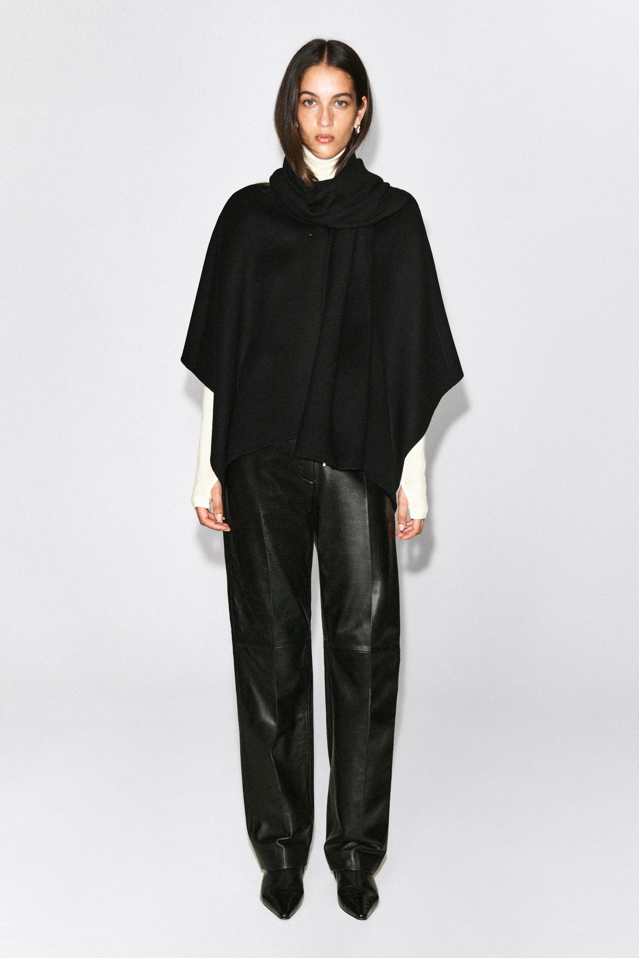 A black cashmere scarf is being worn by the model, draped over the shoulders and wrapped around the neck. The oversized scarf is layered over a cream turtleneck sweater and styled with black leather trousers.