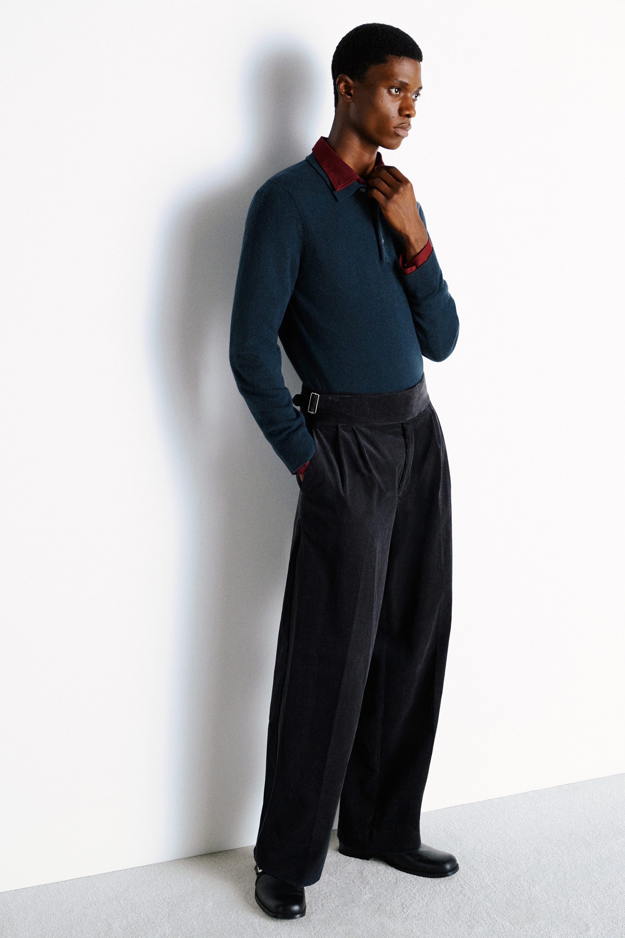 Shown in a three-quarter front view, the teal cashmere polo jumper features a classic collar layered over a burgundy collared shirt. The jumper is tucked into dark gray corduroy relaxed-fit wide-leg pants, highlighting a high-waisted silhouette with front pleats and a side-buckle belt detail. Shown in a three-quarter front view, the teal cashmere polo jumper features a classic collar layered over a burgundy collared shirt. The jumper is tucked into dark gray corduroy relaxed-fit wide-leg pants, highlighting a high-waisted silhouette with front pleats and a side-buckle belt detail.