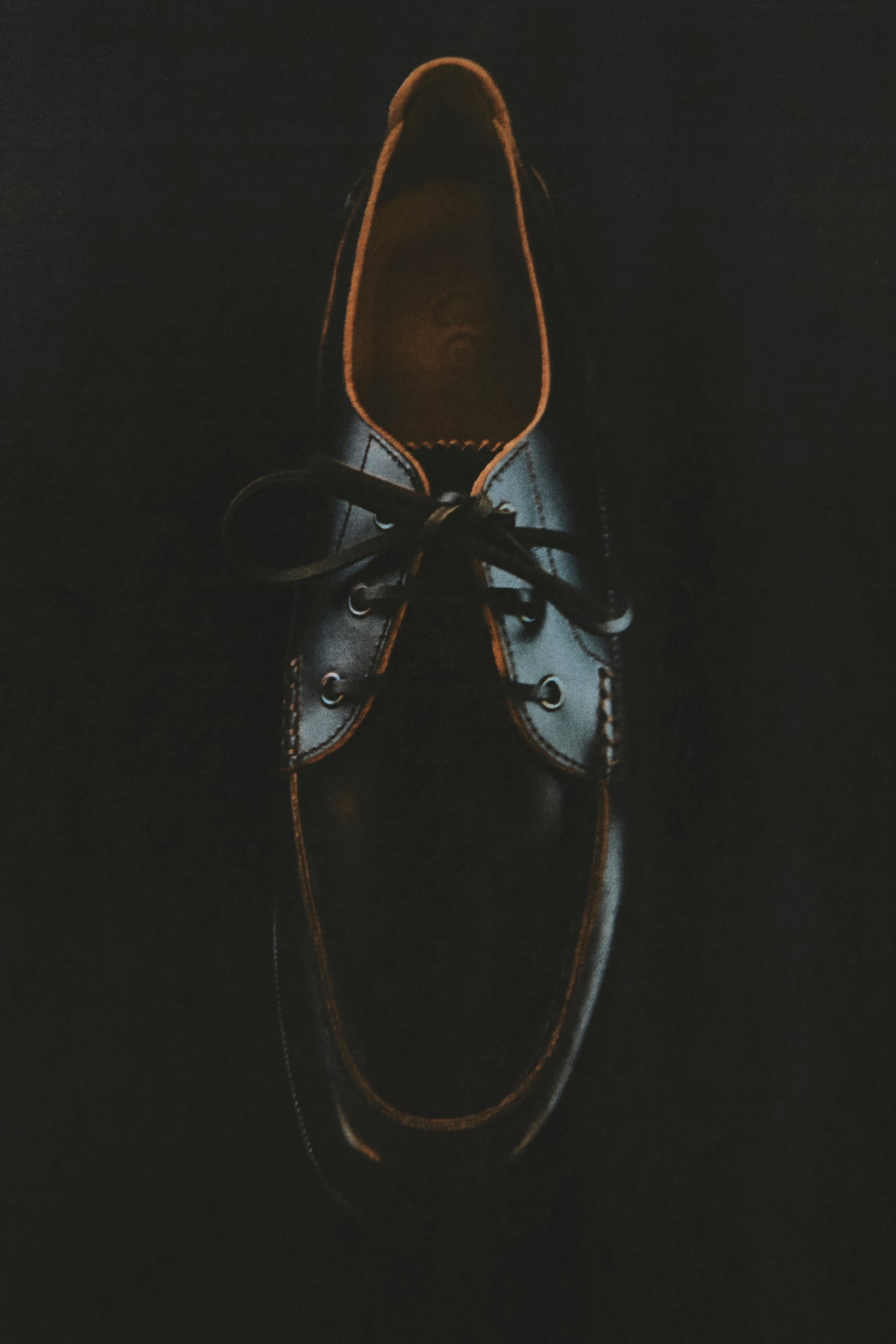 Een donker leren mocassin-stijl schoen wordt gepresenteerd vanuit een bovenaanzicht, met contrasterende tan stiksels rond de neus en een zichtbare tan binnenvoering. Donkere veters zijn door metalen oogjes op de wreef geregen. - 1