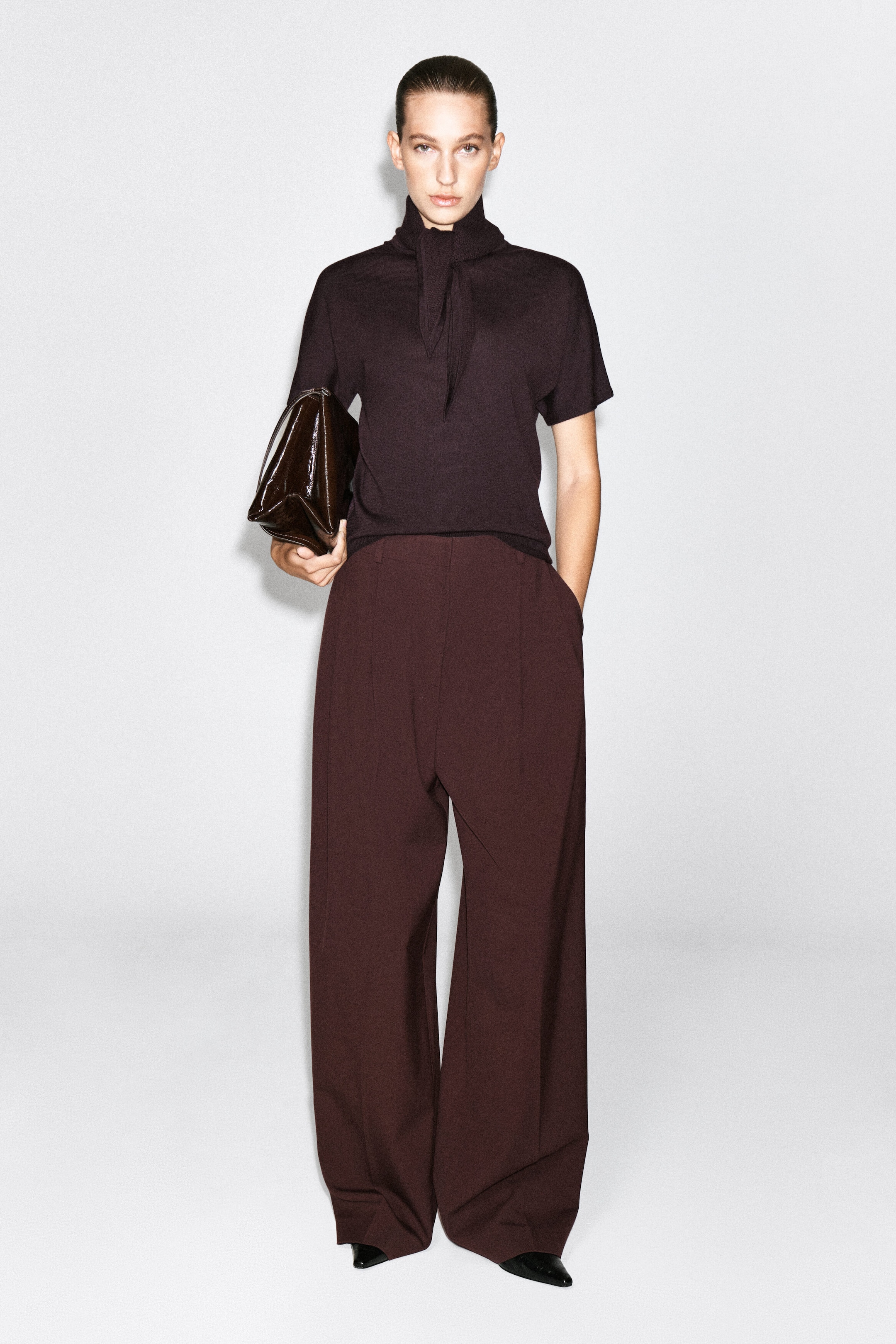 A dark plum-colored merino wool-blend T-shirt featuring a high neck with an integrated, draped scarf detail is worn by the model. The short-sleeved knit top is paired with matching high-waisted, wide-leg trousers.