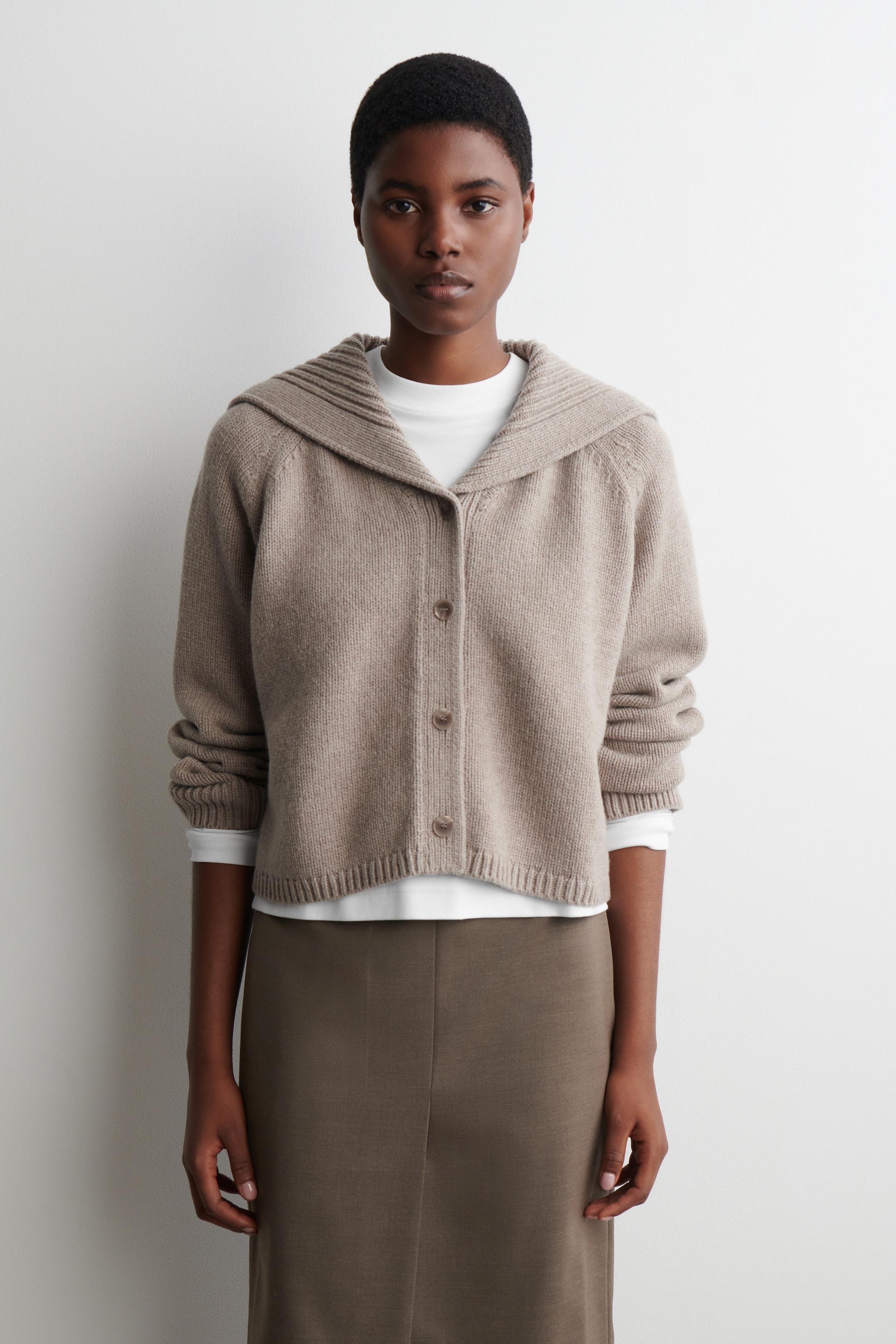 Visualizza immagine più grande: Un cardigan lavorato a maglia in grigio mélange, caratterizzato da una forma squadrata, un colletto a coste stile marinaio e tre bottoni fissati sul davanti, viene indossato. La modella sta stratificando il cardigan sopra una maglia bianca a maniche lunghe e lo sta abbinando a una gonna verde oliva scura.
