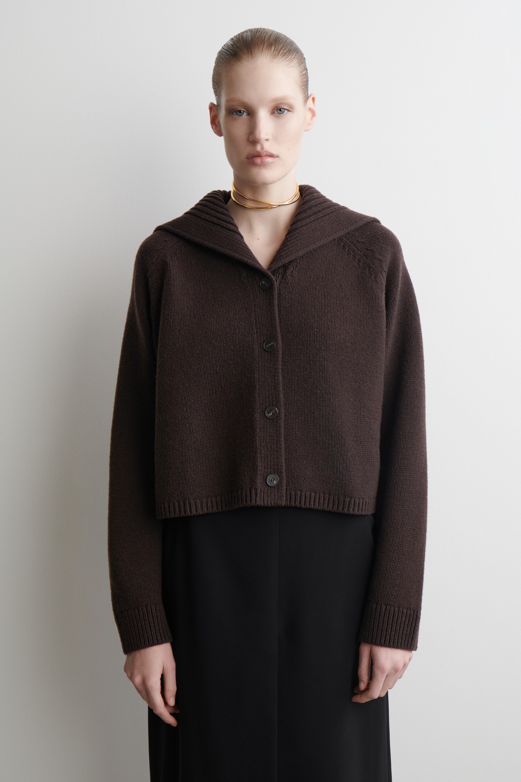 Visualizza immagine più grande: Un cardigan lavorato a maglia di colore marrone scuro è presentato frontalmente, con un colletto con cappuccio, maniche raglan e una lunghezza accorciata. È chiuso sul davanti con quattro bottoni scuri e rifinito con polsini e orlo a coste, indossato sopra un indumento nero.