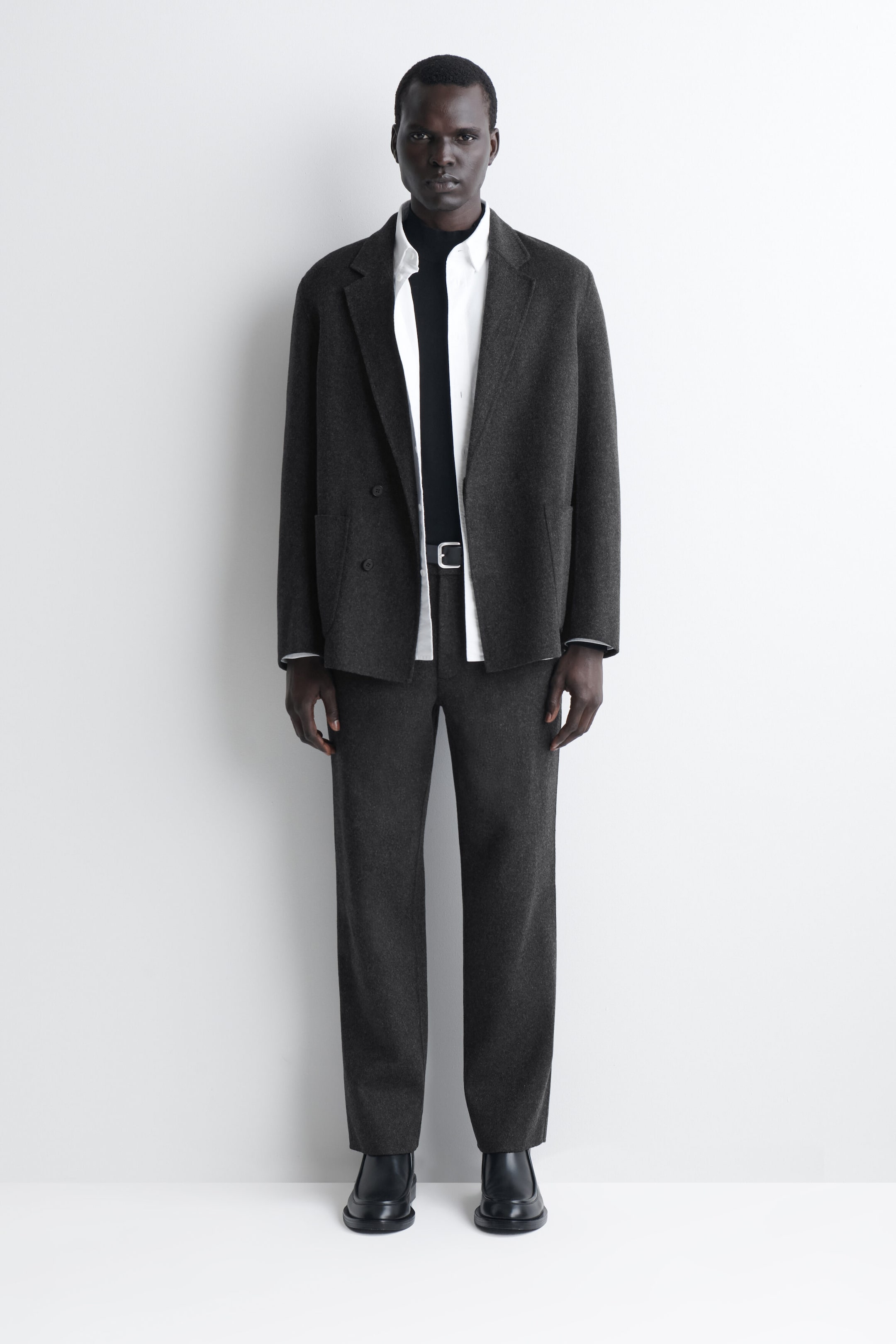 A model is wearing these dark grey, relaxed-fit trousers from a front-facing angle, showcasing their unstructured straight-leg shape and soft drape. The trousers are styled with a black leather belt, a matching double-faced blazer, an open white shirt over a black high-neck top, and black leather boots. A model is wearing these dark grey, relaxed-fit trousers from a front-facing angle, showcasing their unstructured straight-leg shape and soft drape. The trousers are styled with a black leather belt, a matching double-faced blazer, an open white shirt over a black high-neck top, and black leather boots.