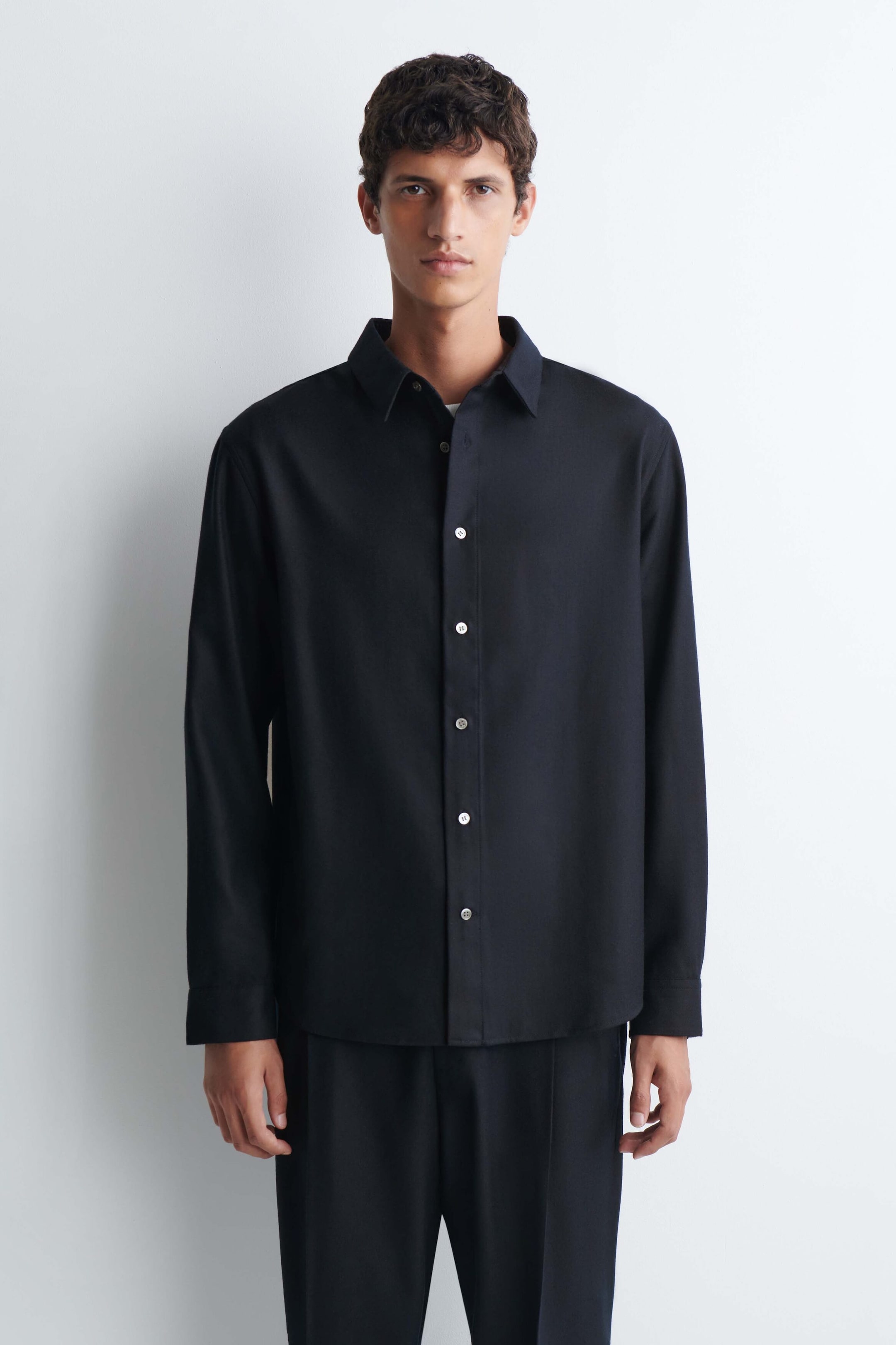 Ein lässiges Hemd aus Wollflanell in Schwarz wird vom Model getragen und verfügt über einen spitzen Kragen, lange Ärmel und kontrastierende helle Knöpfe auf der Vorderseite. Das Hemd ist mit einer passenden dunklen Hose kombiniert. Ein lässiges Hemd aus Wollflanell in Schwarz wird vom Model getragen und verfügt über einen spitzen Kragen, lange Ärmel und kontrastierende helle Knöpfe auf der Vorderseite. Das Hemd ist mit einer passenden dunklen Hose kombiniert.