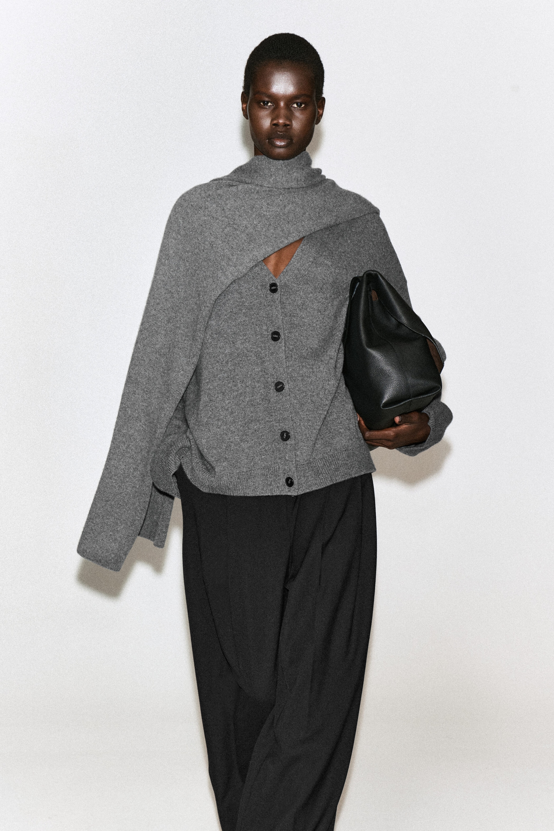 Un cardigan con scollo a V in puro cashmere grigio con bottoni scuri è indossato dalla modella, abbinato a un coprispalle in maglia grigio coordinato drappeggiato su spalla e collo. Il cardigan è abbinato a pantaloni neri a gamba larga, mentre una borsa in pelle nera è portata sotto il braccio della modella. Un cardigan con scollo a V in puro cashmere grigio con bottoni scuri è indossato dalla modella, abbinato a un coprispalle in maglia grigio coordinato drappeggiato su spalla e collo. Il cardigan è abbinato a pantaloni neri a gamba larga, mentre una borsa in pelle nera è portata sotto il braccio della modella.