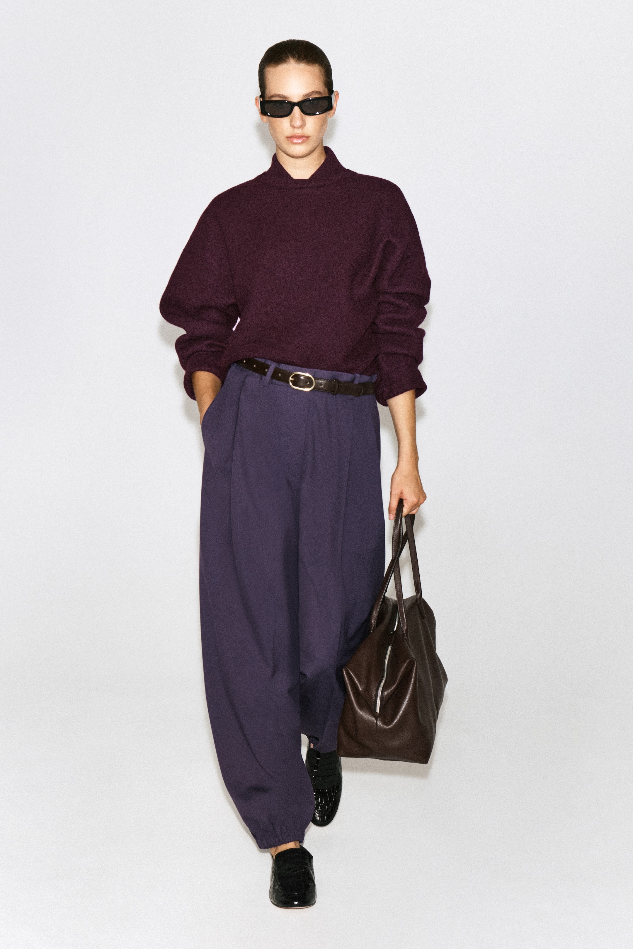 Agrandir l'image: Le mannequin porte un pantalon ballon plissé dans une teinte violet foncé, doté d’une taille haute avec des plis sur le devant et une jambe volumineuse resserrée à la cheville. Le pantalon est associé à un pull bordeaux rentré, à une ceinture marron foncé et à des mocassins en cuir noirs.