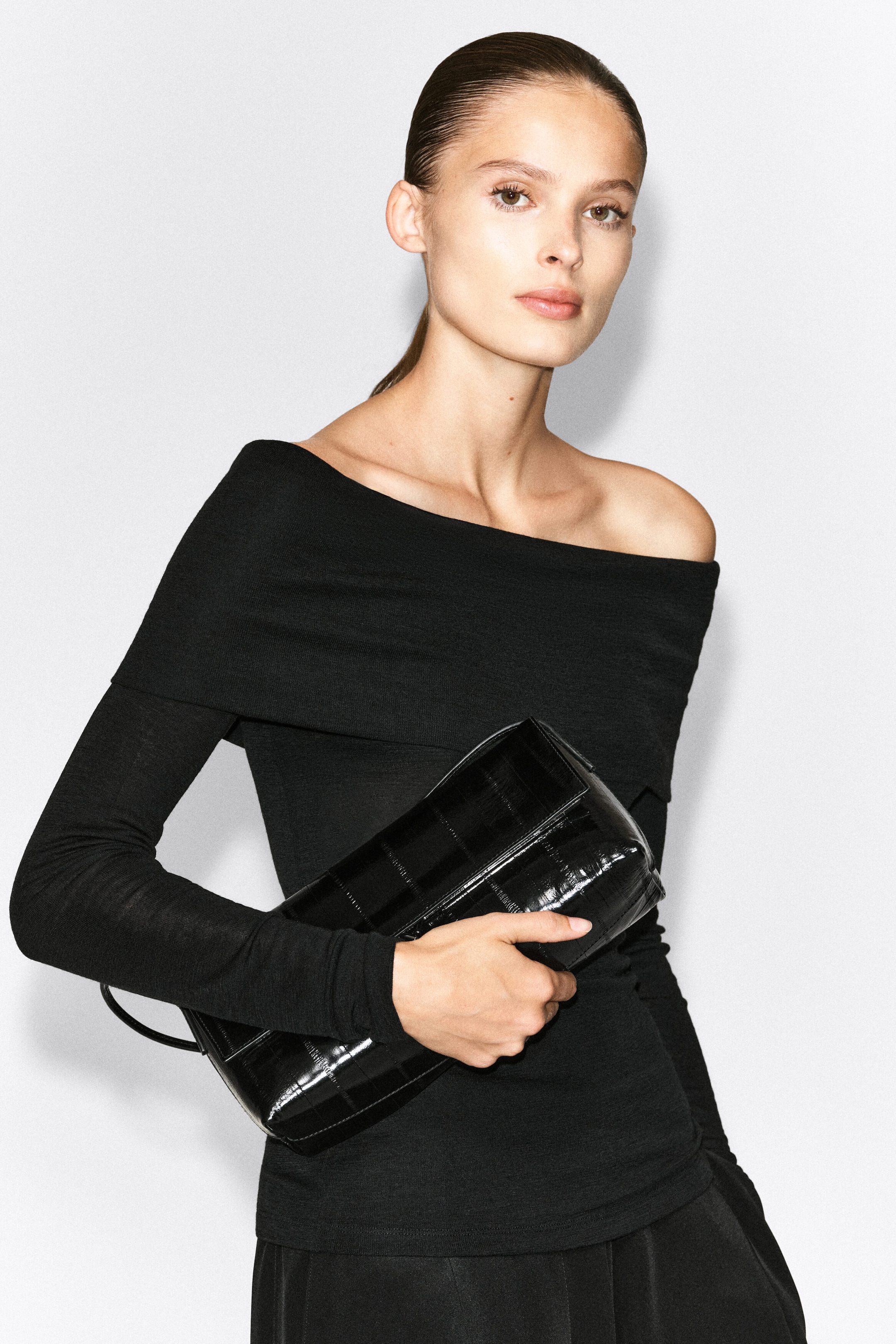A black off-the-shoulder merino wool top featuring a wide, draped neckline and long sleeves is worn by the model. The model is holding a black crocodile-embossed clutch bag.