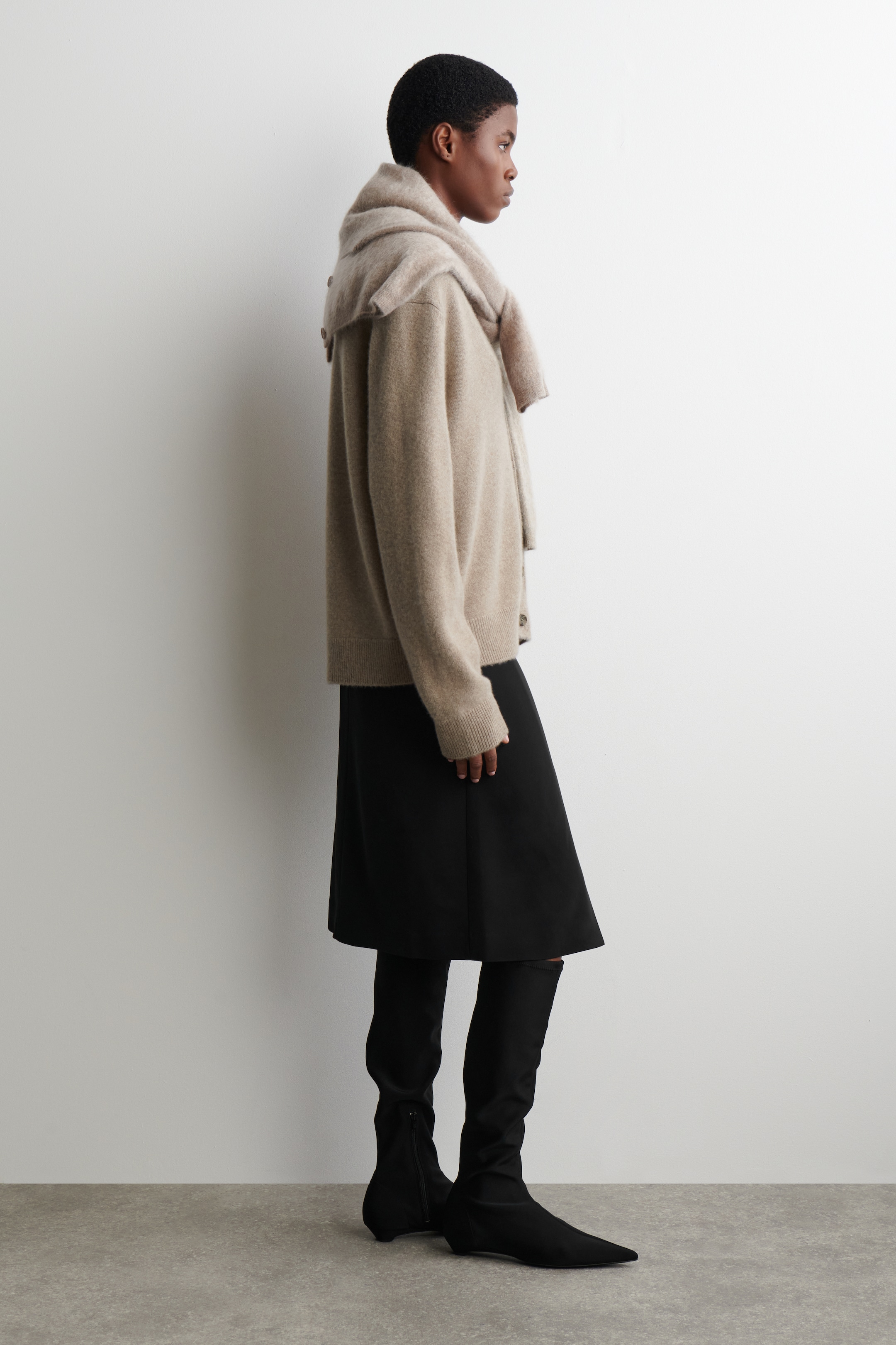 View larger image: A beige cardigan, featuring ribbed cuffs and a relaxed, open-front design, is being presented from a side profile. A matching beige scarf is being draped over its shoulders, and it is being worn over a black knee-length skirt and black pointed-toe knee-high boots.