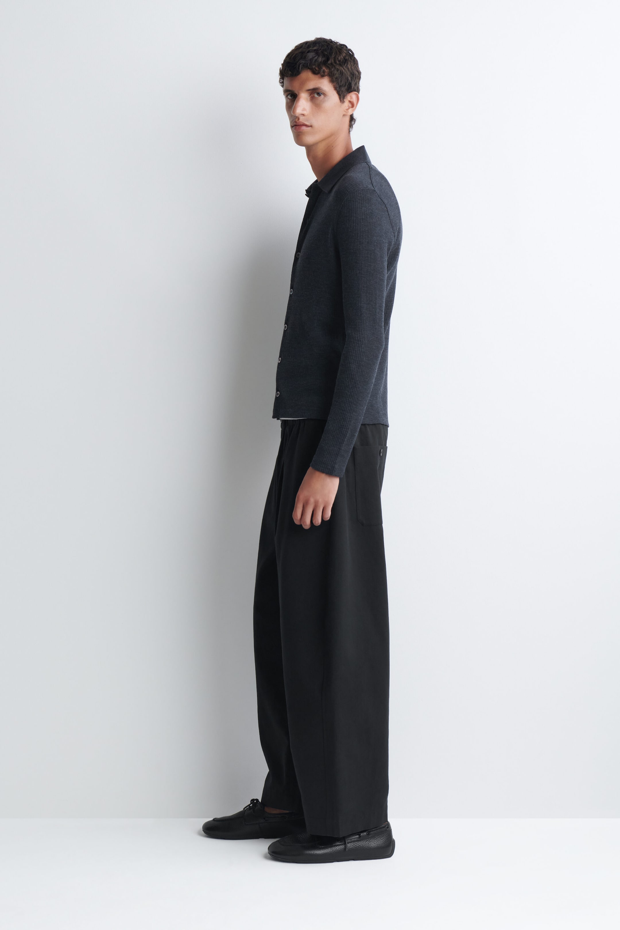 A dark grey, long-sleeved, ribbed knit cardigan with a collar is being worn by the model, paired with wide-leg black trousers and black leather loafers.