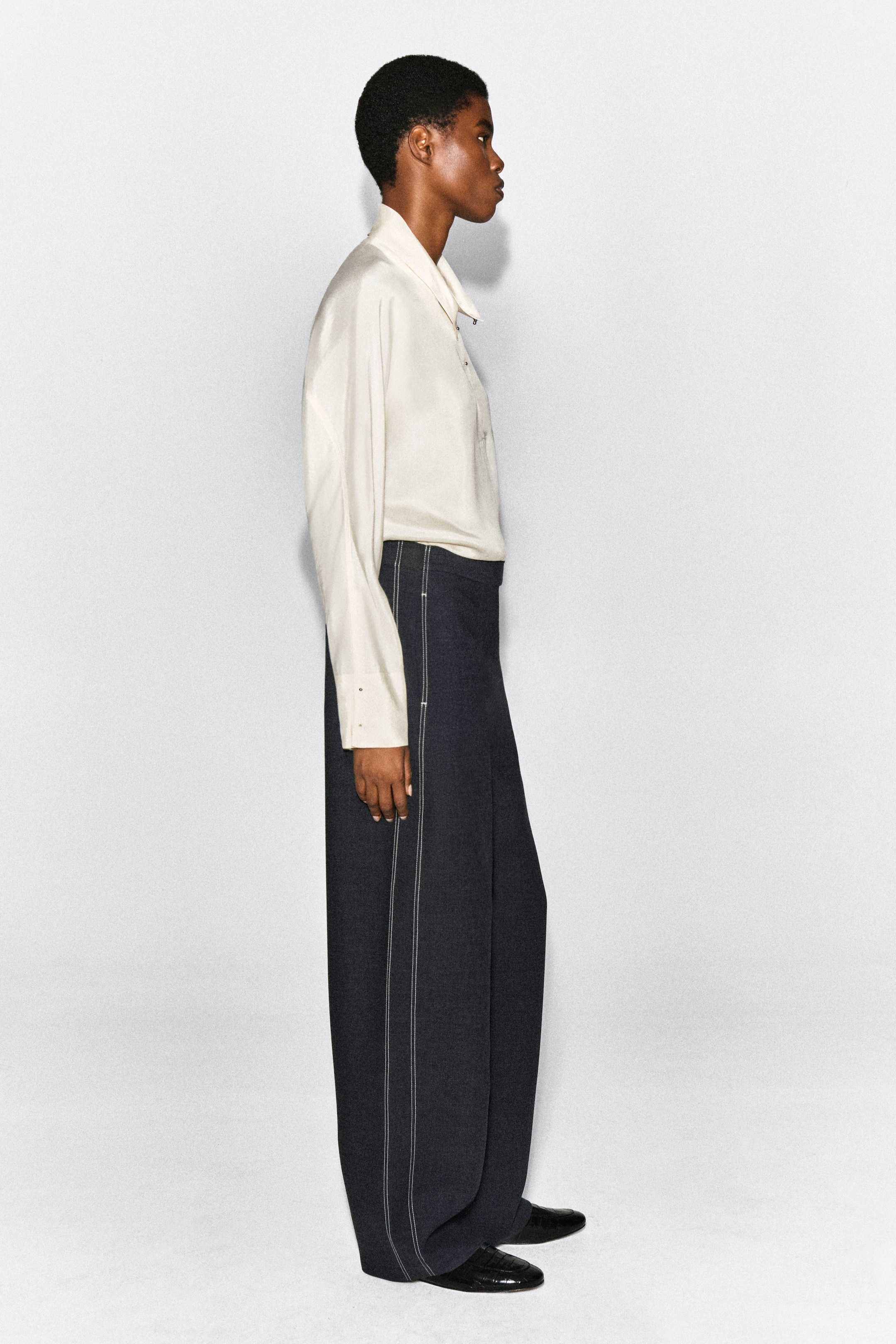 Agrandir l'image: Un pantalon barrel à surpiqûres dans un tissu gris foncé est porté par le mannequin, mettant en valeur une silhouette arrondie et des surpiqûres blanches contrastantes bien visibles le long de la couture latérale. Ce pantalon taille haute est associé à une blouse crème à manches longues rentrée et à des chaussures noires.