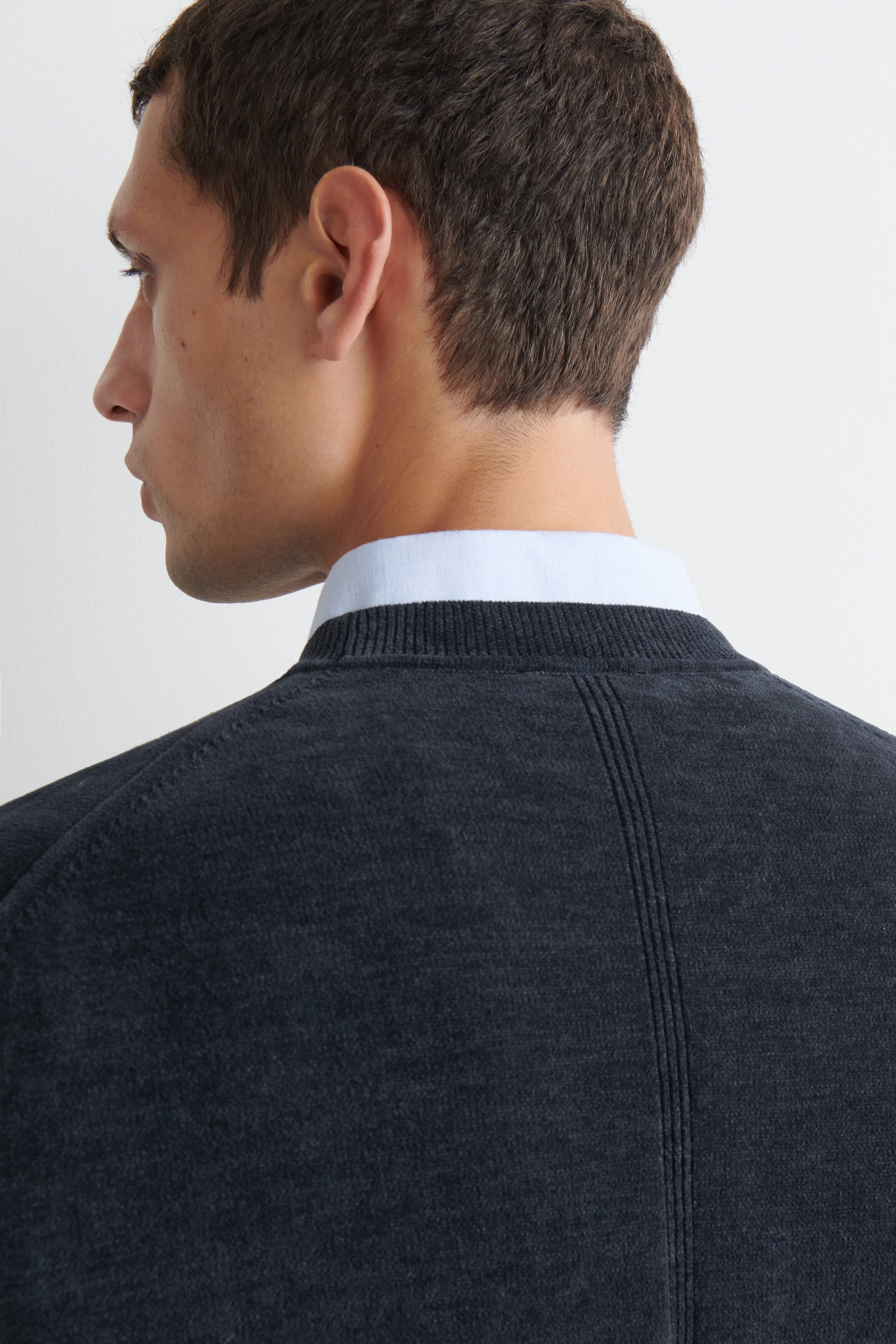 The cotton-chenille crewneck sweater is shown from the back, featuring a dark gray fine-knit texture, a ribbed crew neckline, and a vertical seam running down the center back. The sweater is being worn layered over a light blue collared shirt. The cotton-chenille crewneck sweater is shown from the back, featuring a dark gray fine-knit texture, a ribbed crew neckline, and a vertical seam running down the center back. The sweater is being worn layered over a light blue collared shirt.