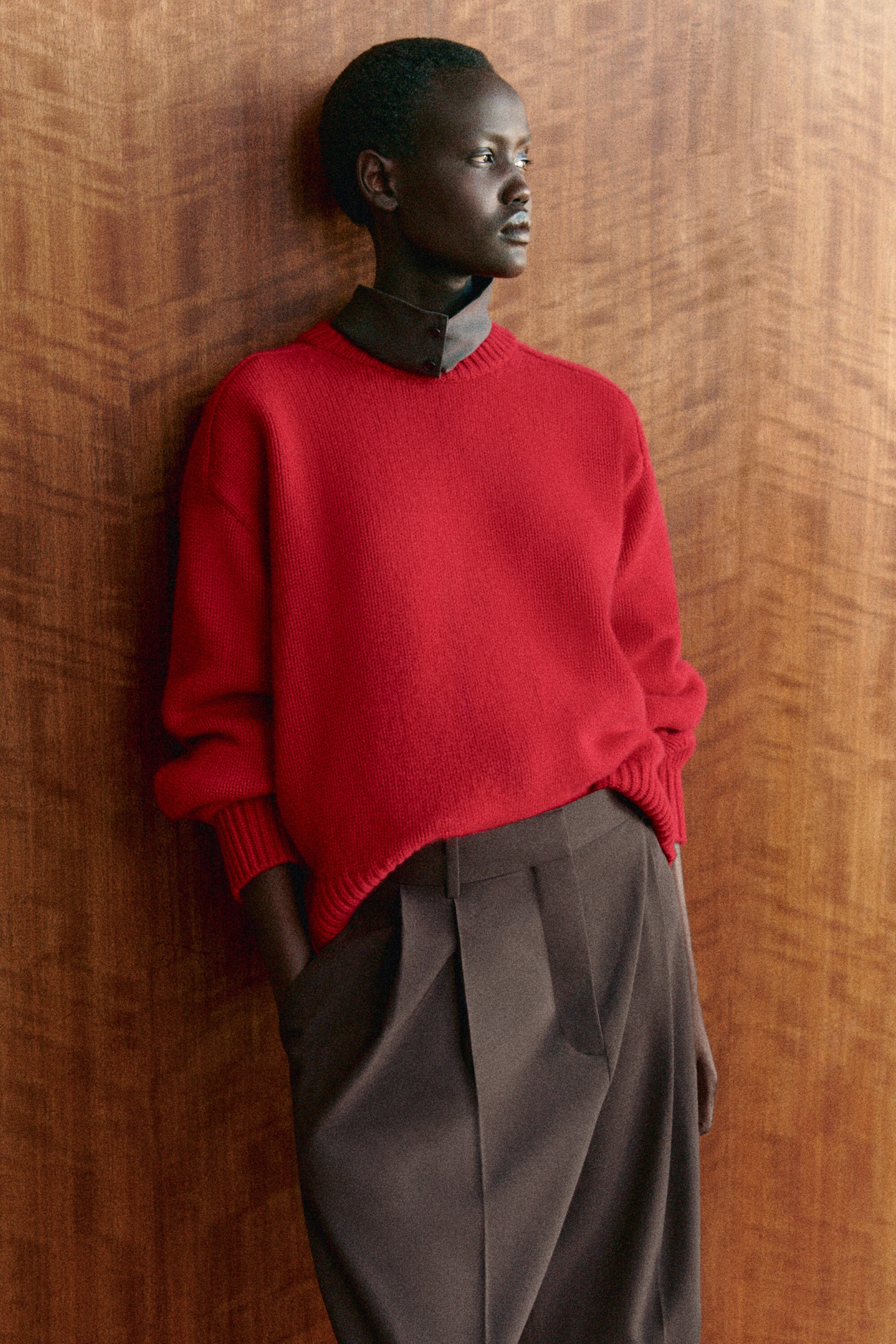 A red, rounded wool crew-neck sweater is shown from the front, featuring a relaxed, oversized fit with dropped shoulders and ribbed detailing at the neckline, cuffs, and hem. The sweater is styled layered over a dark grey collared shirt and partially tucked into brown pleated trousers. A red, rounded wool crew-neck sweater is shown from the front, featuring a relaxed, oversized fit with dropped shoulders and ribbed detailing at the neckline, cuffs, and hem. The sweater is styled layered over a dark grey collared shirt and partially tucked into brown pleated trousers.