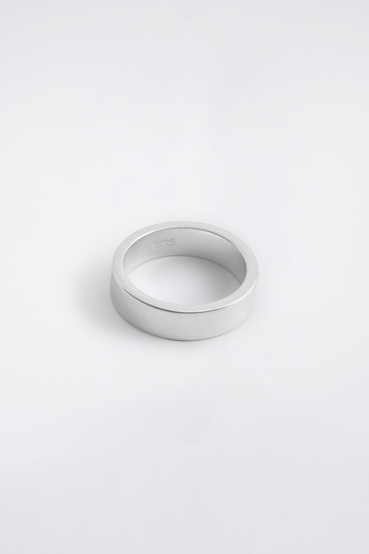 A silver-colored band ring with a brushed matte finish is resting on a light surface, showing its clean, flat-edged profile from a three-quarter angle. A small logo is engraved on the inside of the band.