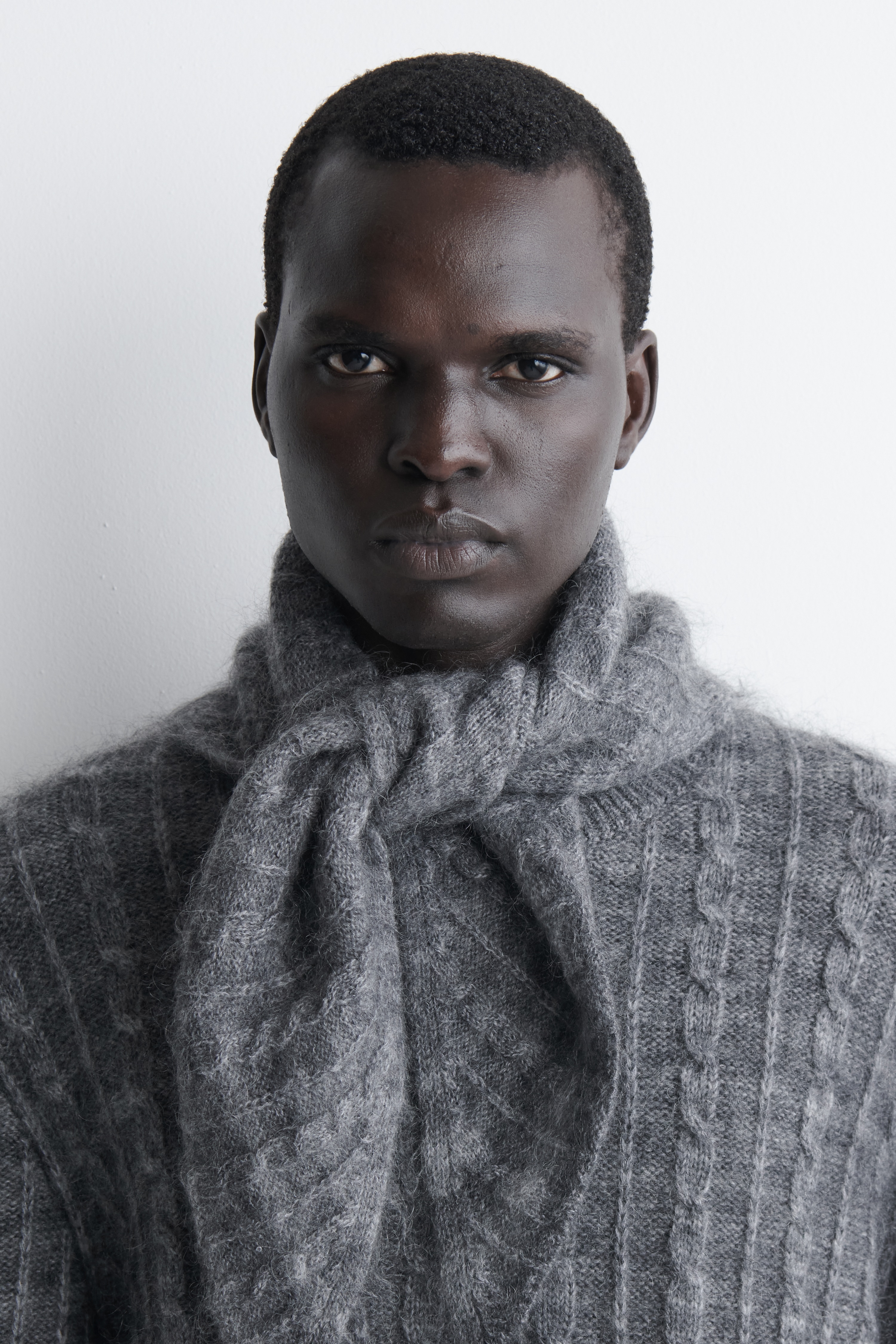 En mÃ¶rkgrÃ¥ rutig kabelstickad triangelscarf visas framifrÃ¥n, med en mjuk, borstad stickad struktur med subtila vertikala kabeldetaljer och rutmÃ¶nster. Scarfen Ã¤r knuten i en lÃ¶s knut i mitten, med sina avsmalnande sidÃ¤ndar och triangulÃ¤ra spets som faller nedÃ¥t.