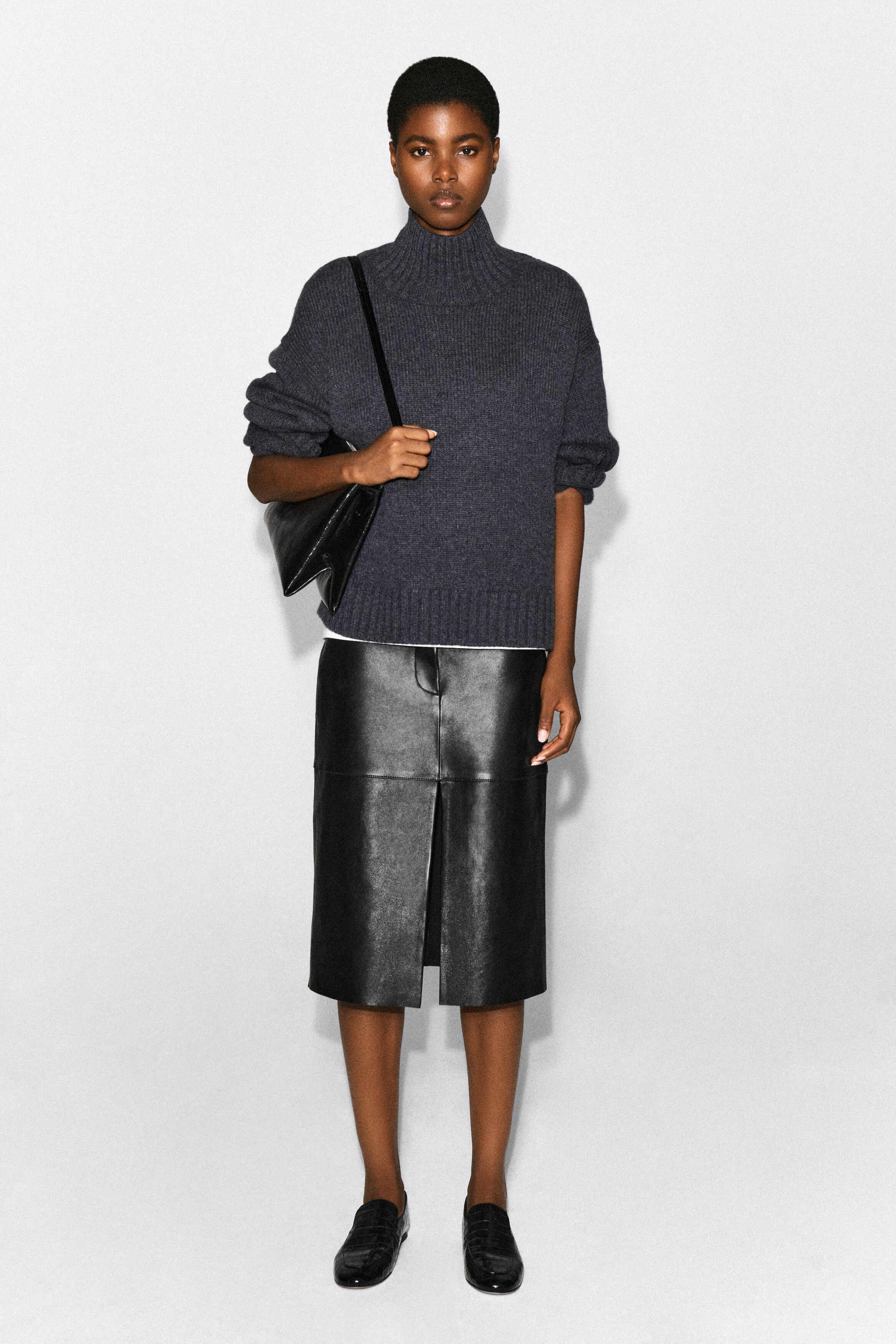 A chunky charcoal-grey cashmere jumper featuring a ribbed funnel neck, hem and cuffs is being worn by the model. The jumper is styled with a black leather midi skirt and a black leather shoulder bag held under the model’s arm. A chunky charcoal-grey cashmere jumper featuring a ribbed funnel neck, hem and cuffs is being worn by the model. The jumper is styled with a black leather midi skirt and a black leather shoulder bag held under the model’s arm.