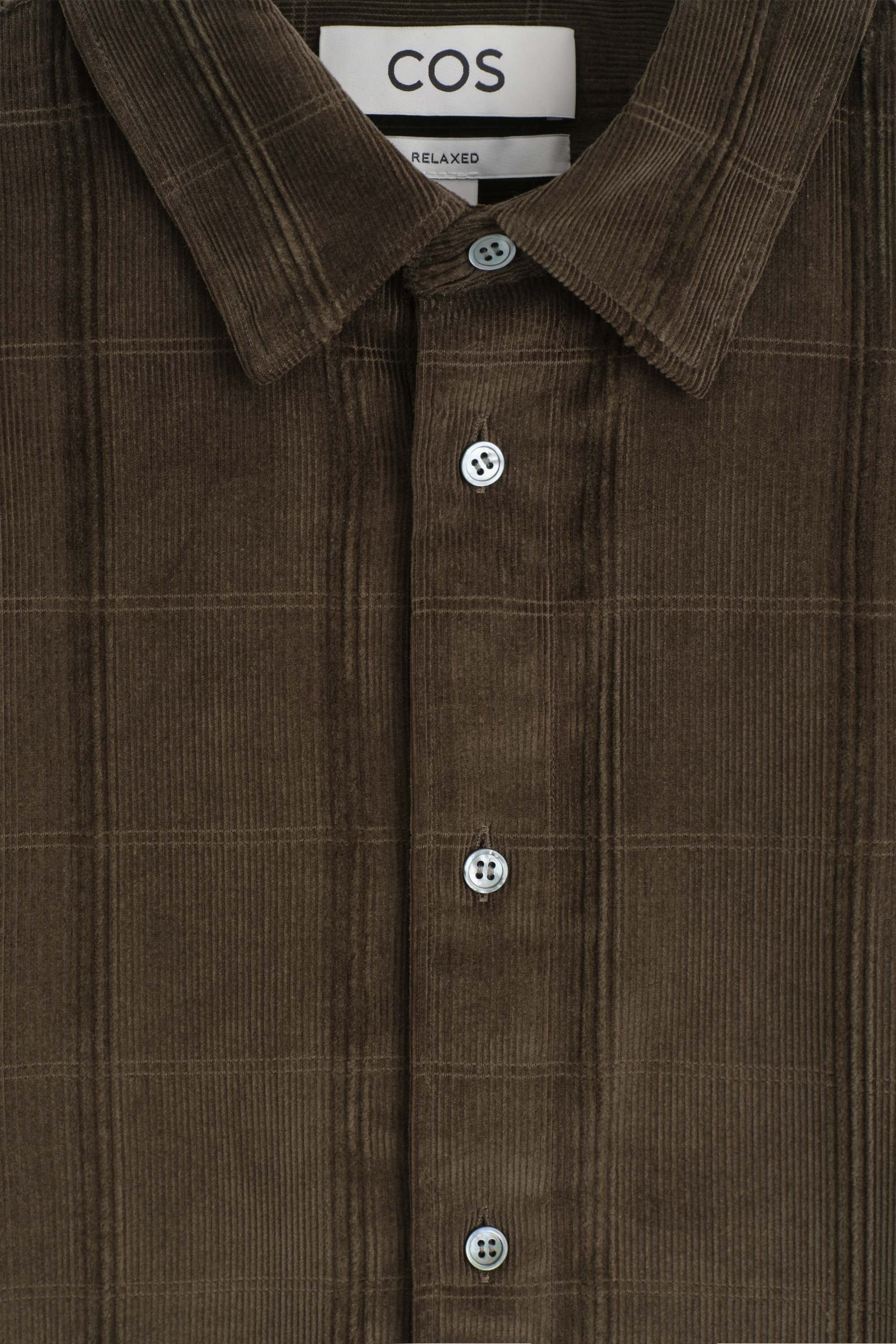 The Relaxed Corduroy Shirt is shown in a close-up front view, displaying a dark brown corduroy fabric with a subtle grid pattern. The shirt features a classic pointed collar, a front placket with light grey buttons, and a white brand label at the inner neckline. The Relaxed Corduroy Shirt is shown in a close-up front view, displaying a dark brown corduroy fabric with a subtle grid pattern. The shirt features a classic pointed collar, a front placket with light grey buttons, and a white brand label at the inner neckline.