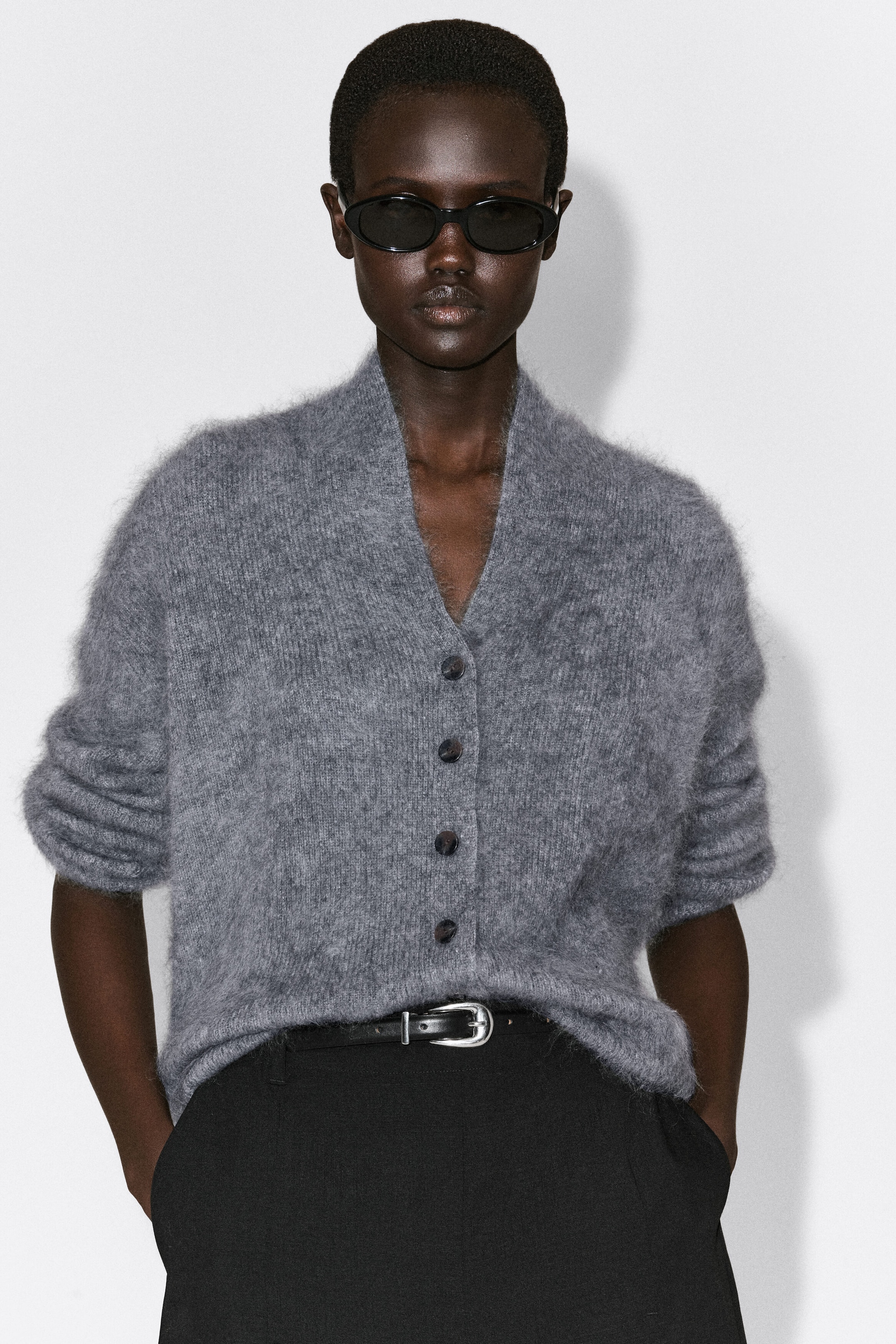 The draped brushed merino wool cardigan is facing front, showcasing a fuzzy gray knit texture, a deep V-neckline, and a front button closure. The relaxed-fit cardigan is being worn tucked into black trousers with a belt, featuring its long sleeves pushed up to the elbows.