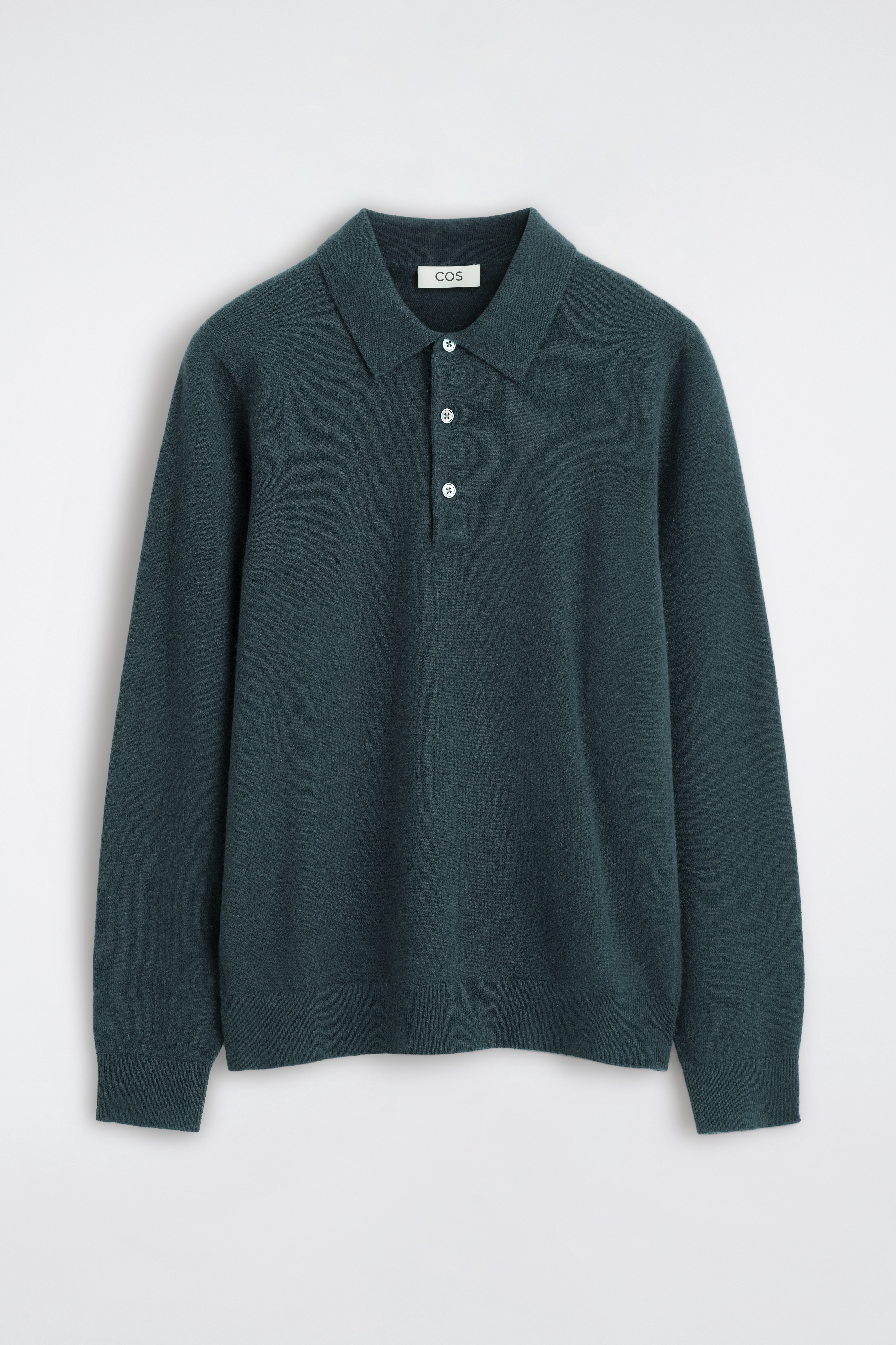 Un maglione polo in cashmere verde scuro, con colletto classico, abbottonatura a tre bottoni con bottoni chiari a contrasto e bordi a coste, indossato dalla modella. Il maglione Ã¨ abbinato a pantaloni scuri.