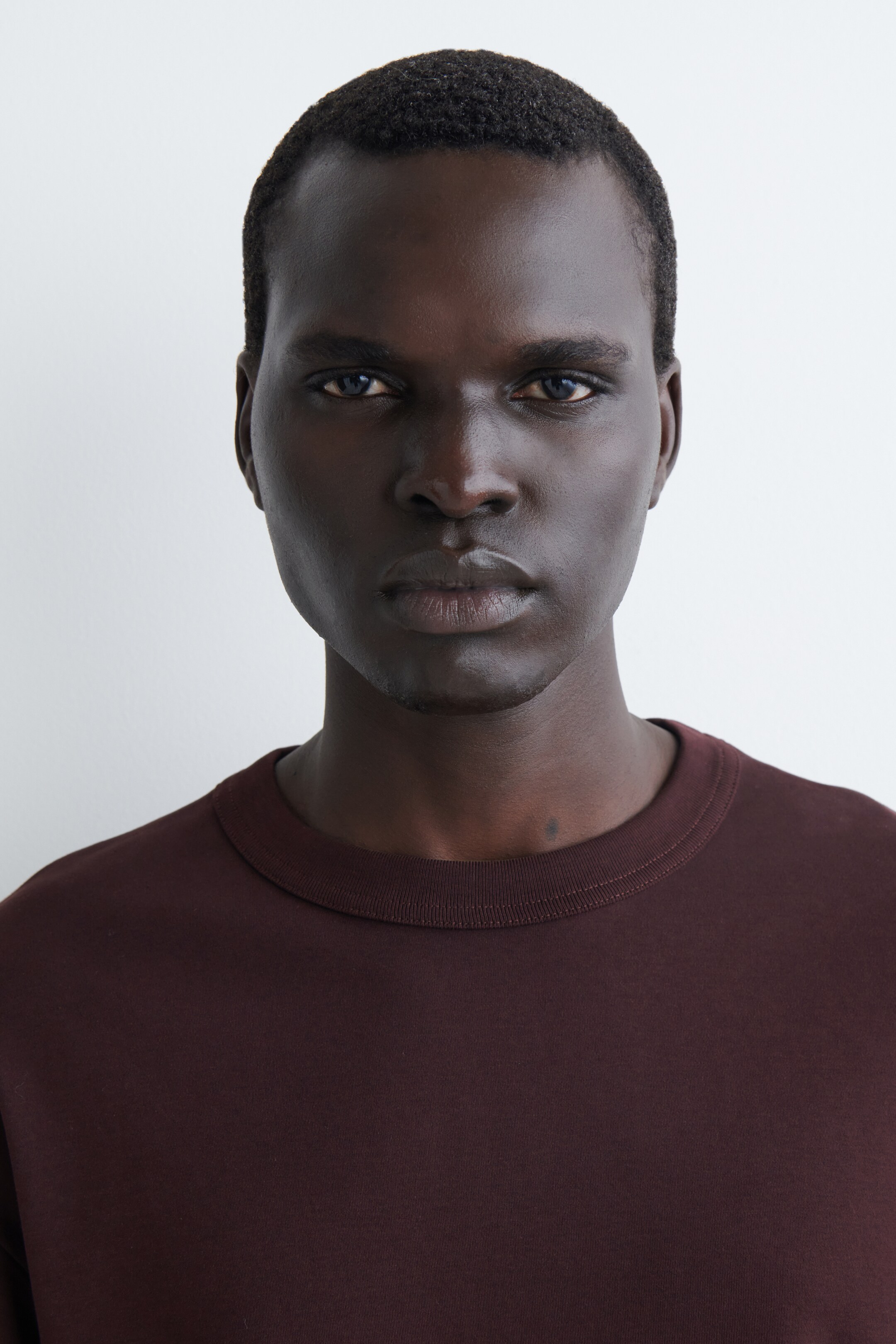 Visualizza immagine più grande: Una t-shirt di colore marrone-rossastro scuro è in mostra, caratterizzata da un classico girocollo con una fascia a costine visibile. Il tessuto solido e liscio mostra una vestibilità rilassata sulle spalle e sulla parte superiore del petto.