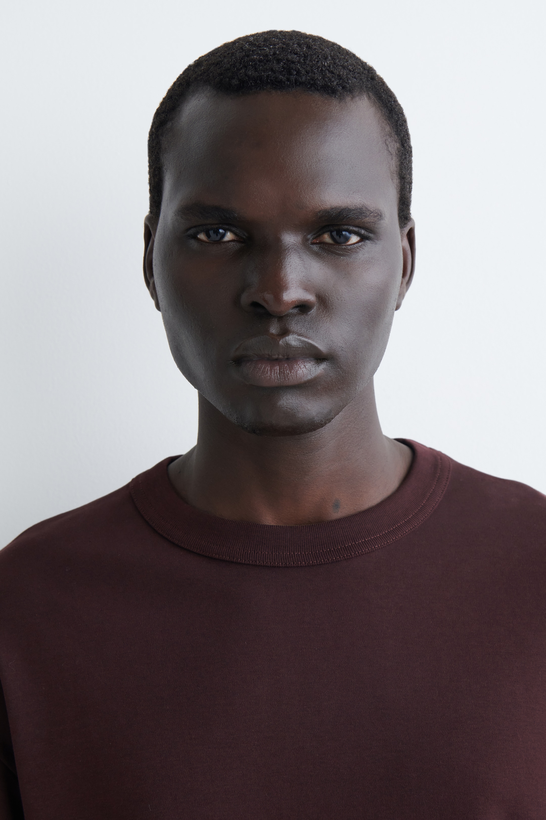 Ver imagen más grande: Una camiseta de color marrón rojizo oscuro está siendo exhibida, con un clásico cuello redondo y una banda de cuello acanalada visible. El tejido sólido y suave muestra un ajuste relajado en los hombros y la parte superior del pecho.