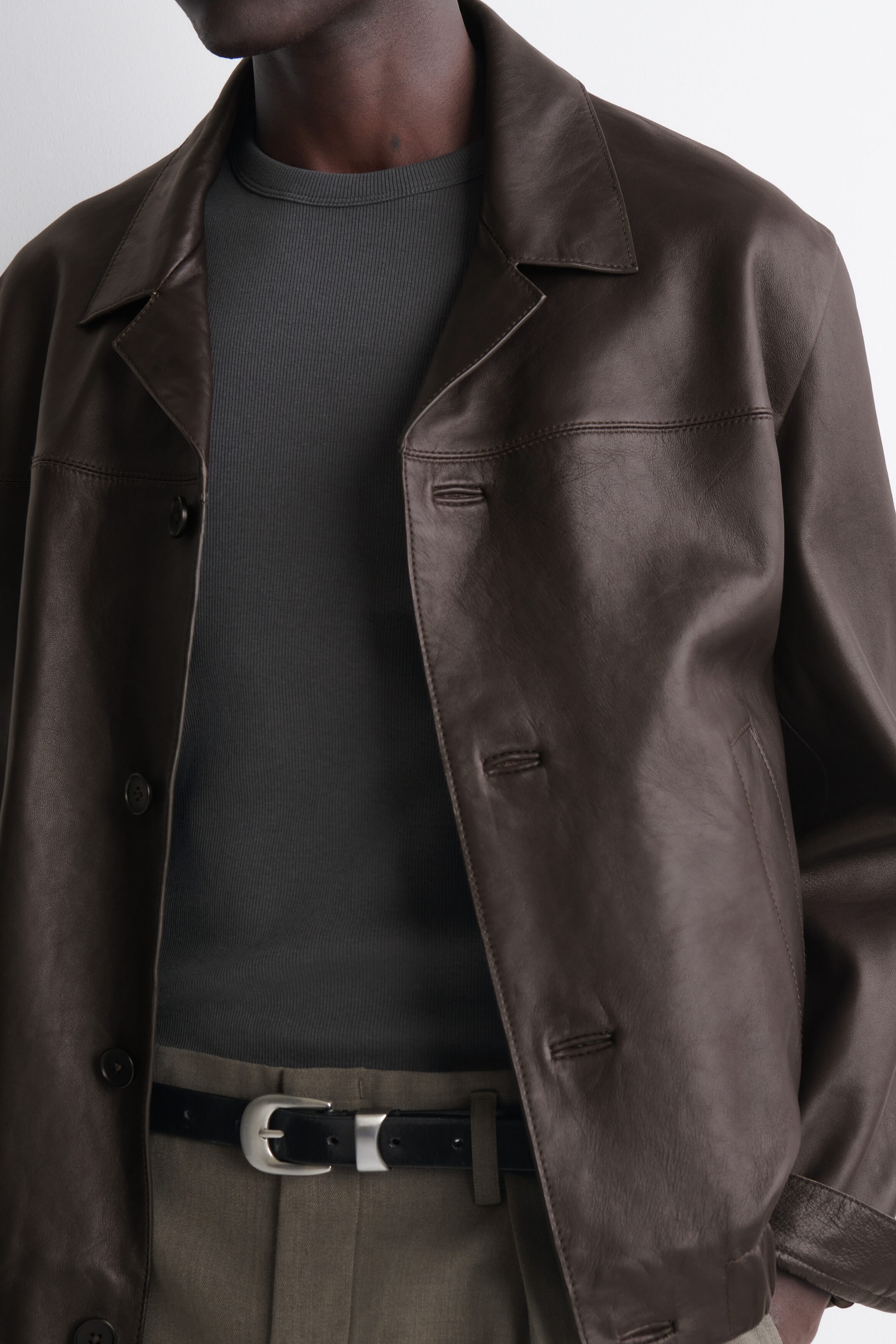 A slim ribbed cotton T-shirt in dark grey features a fine ribbed texture and a classic crew neck. The T-shirt is shown from the front as a base layer beneath an open brown leather jacket, highlighting its close-fitting silhouette.