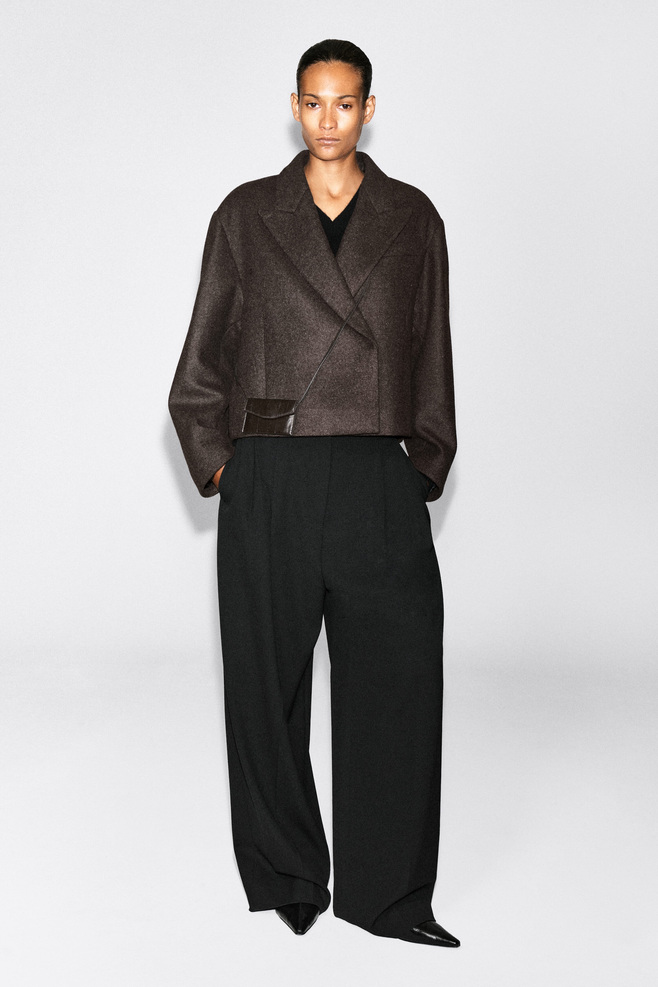 An eel-effect leather cardholder in dark brown is worn crossbody by the model. The small, rectangular accessory features a front flap and a slim strap, layered over a cropped brown wool jacket and wide-leg black trousers. An eel-effect leather cardholder in dark brown is worn crossbody by the model. The small, rectangular accessory features a front flap and a slim strap, layered over a cropped brown wool jacket and wide-leg black trousers.