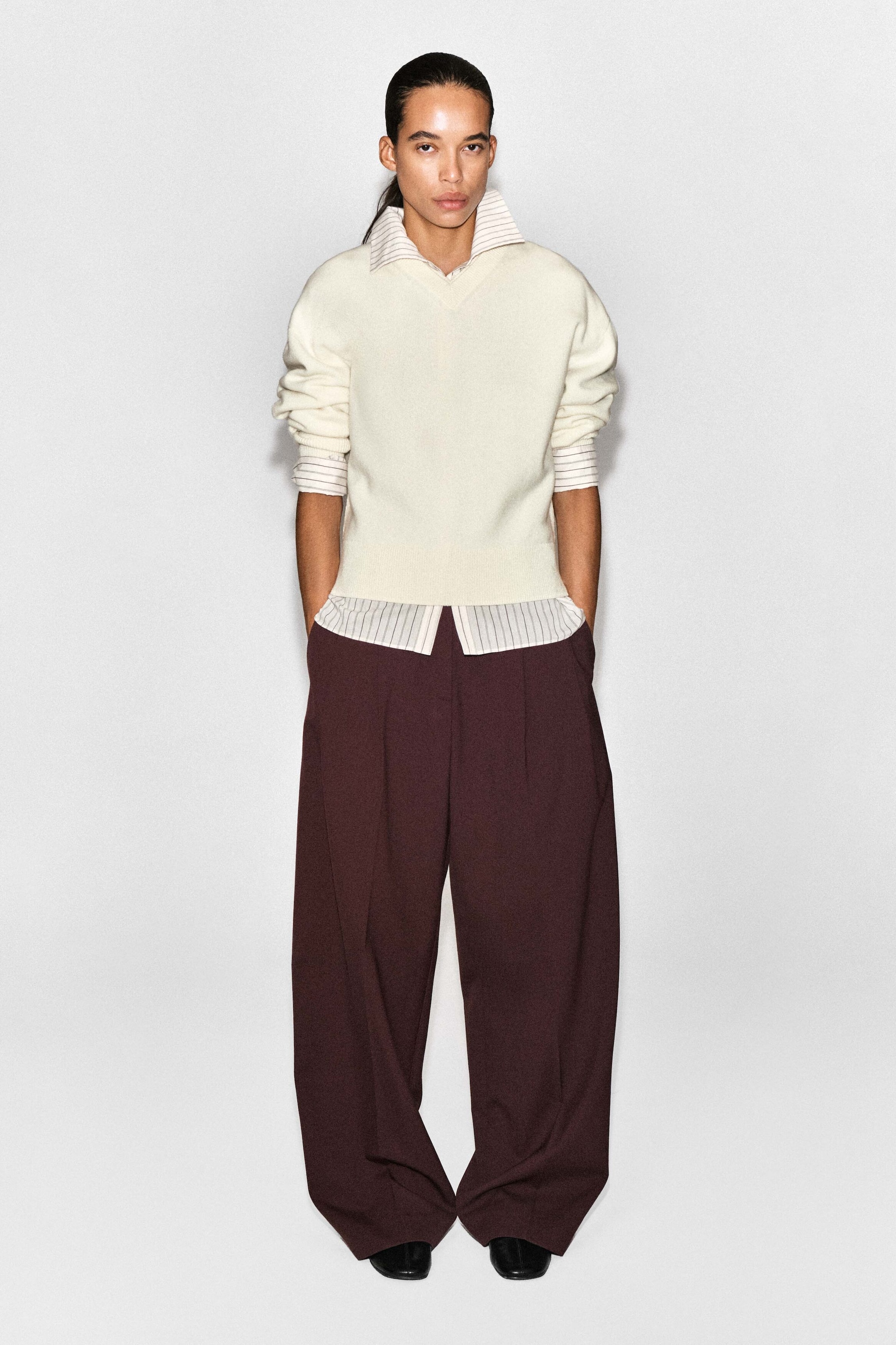 The cream-coloured merino wool layered V-neck jumper is shown from the front, featuring a relaxed silhouette with ribbed cuffs pushed up to the elbows. The jumper drapes over a striped collared shirt peeking out at the neckline, cuffs, and straight hem. The cream-coloured merino wool layered V-neck jumper is shown from the front, featuring a relaxed silhouette with ribbed cuffs pushed up to the elbows. The jumper drapes over a striped collared shirt peeking out at the neckline, cuffs, and straight hem.