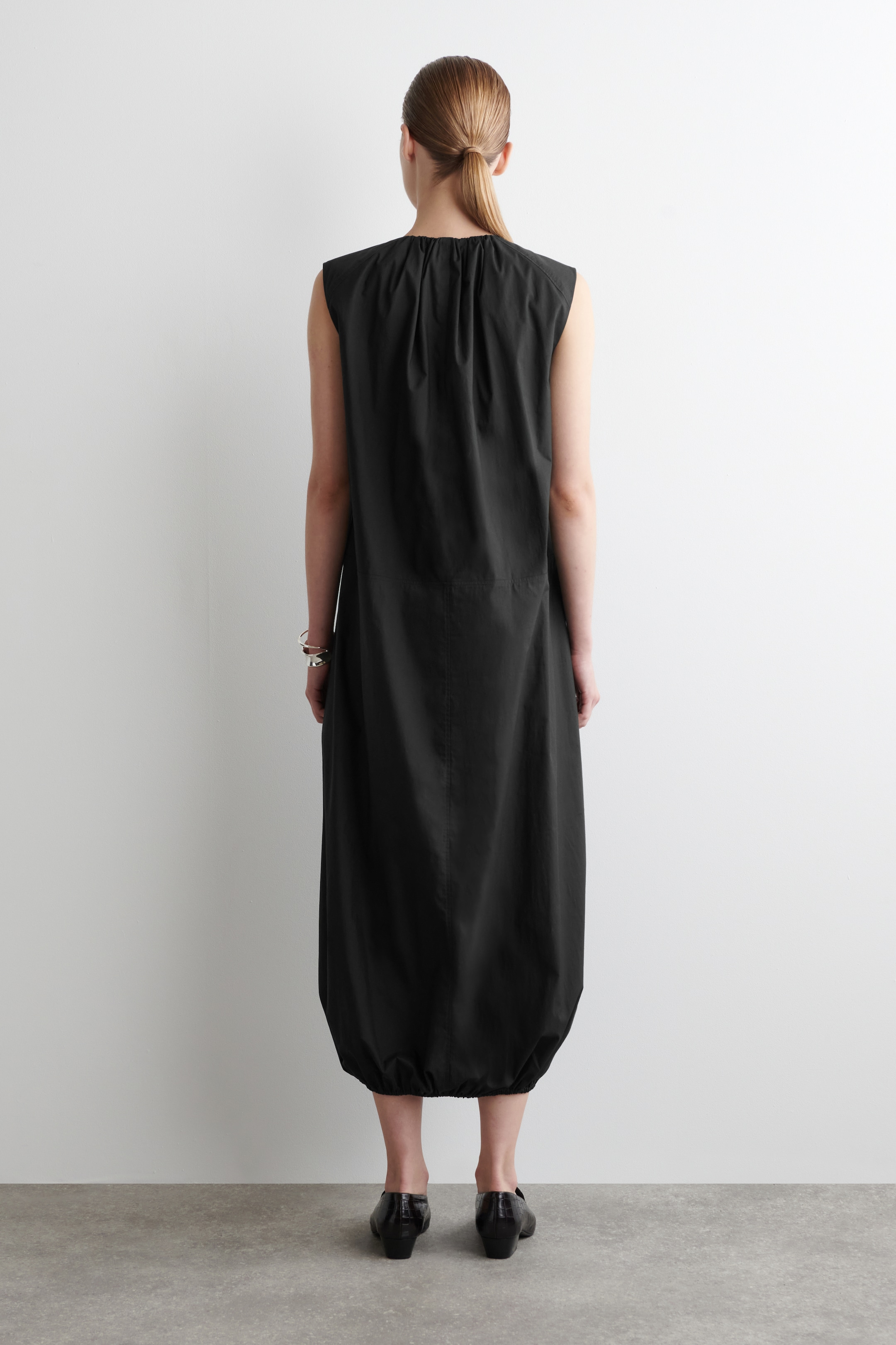 Agrandir l'image: Une robe noire sans manches est présentée de dos, mettant en valeur sa coupe décontractée. La robe présente un détail froncé au niveau du haut du dos et se rétrécit jusqu'à un ourlet style ballon resserré aux chevilles.