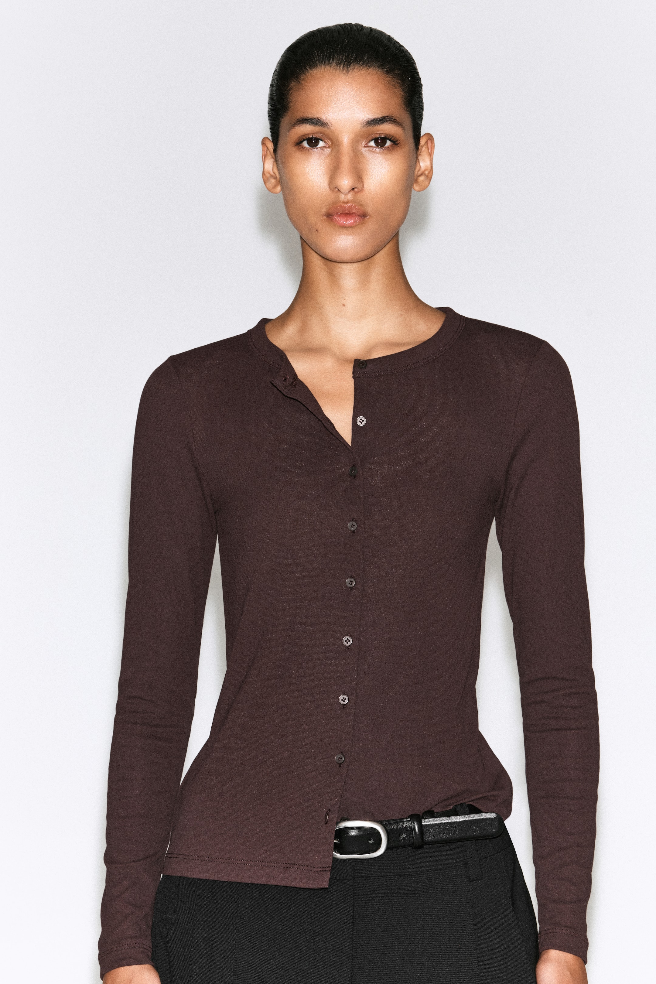 Größeres Bild anzeigen: Dieses dunkelbraune Jersey-Cardigan-Top wird von vorne gezeigt und verfügt über eine schmale Passform, lange Ärmel und einen Rundhalsausschnitt mit Knopfleiste vorne. Das Top wird mit den oberen Knöpfen offen getragen und über einer schwarzen Hose mit Gürtel gestylt, wodurch der leichte Stoff und die figurbetonte Silhouette hervorgehoben werden.