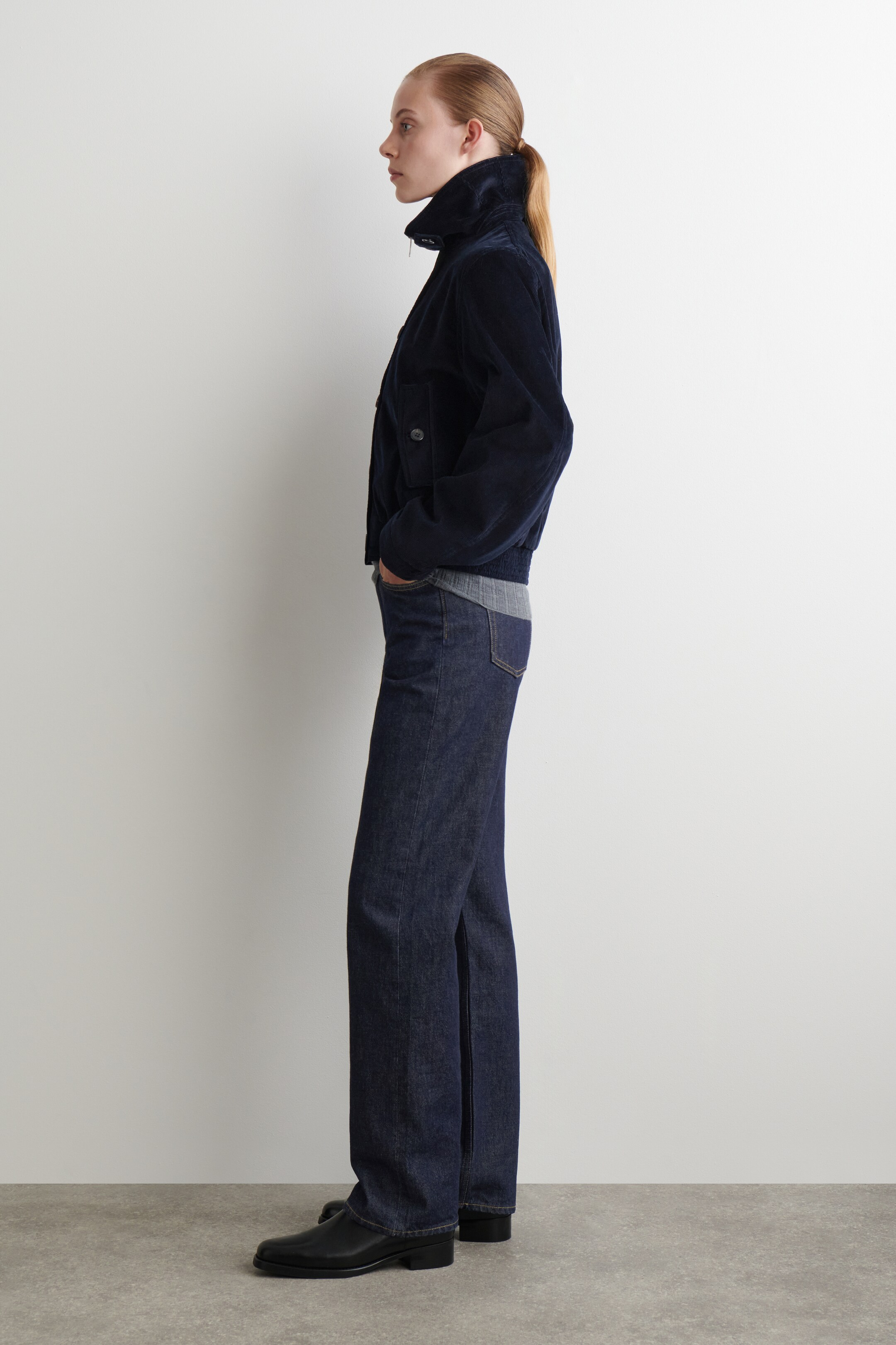 Ver imagem maior: Um blusão bomber de bombazina azul-marinho, com gola alta, dois bolsos com aba e bainha canelada, está a ser usado de perfil. Está a ser combinado com jeans de lavagem escura, de perna reta, um top cinzento de malha canelada e sapatos pretos de salto baixo.