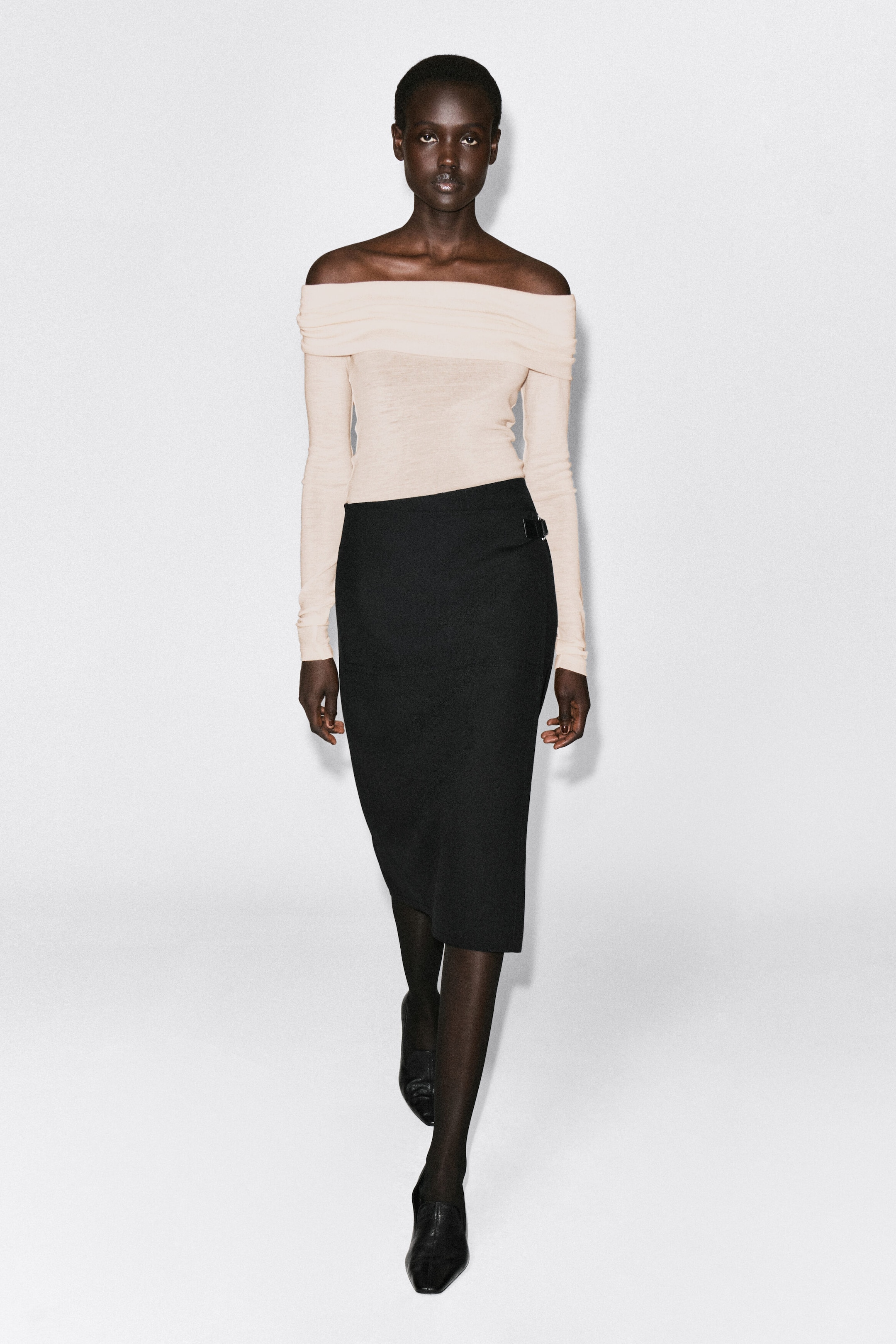 The light beige off-the-shoulder merino wool top is shown from the front, featuring a wide folded neckline and slim-fitting long sleeves. It is tucked into a high-waisted black midi skirt, highlighting its close-fitting silhouette. The light beige off-the-shoulder merino wool top is shown from the front, featuring a wide folded neckline and slim-fitting long sleeves. It is tucked into a high-waisted black midi skirt, highlighting its close-fitting silhouette.