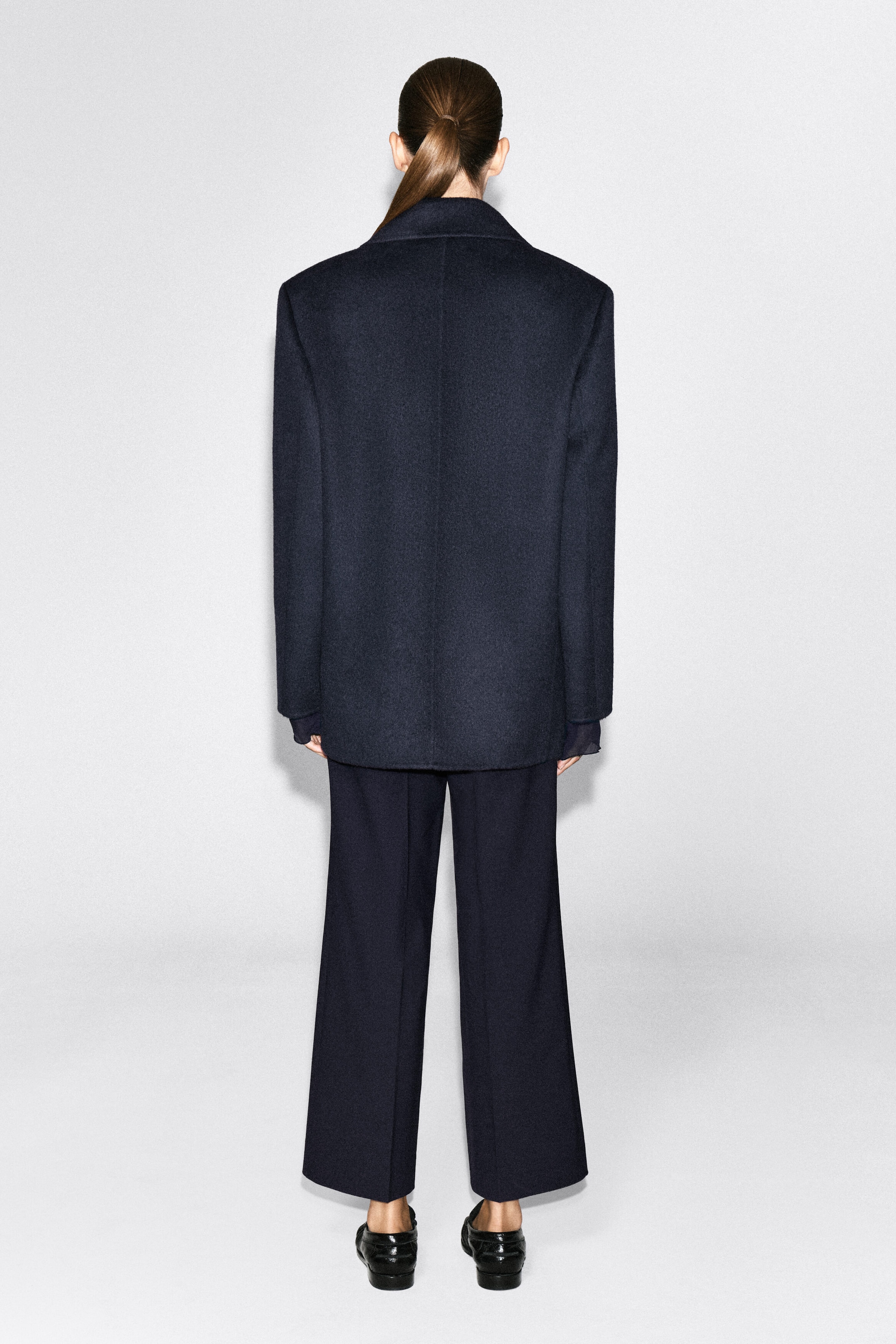 A dark navy wool-blend blazer is being worn by the model, viewed from the back to showcase its structured shoulders, straight fit and central vertical seam. The blazer is paired with matching wide-leg trousers and black leather loafers.
