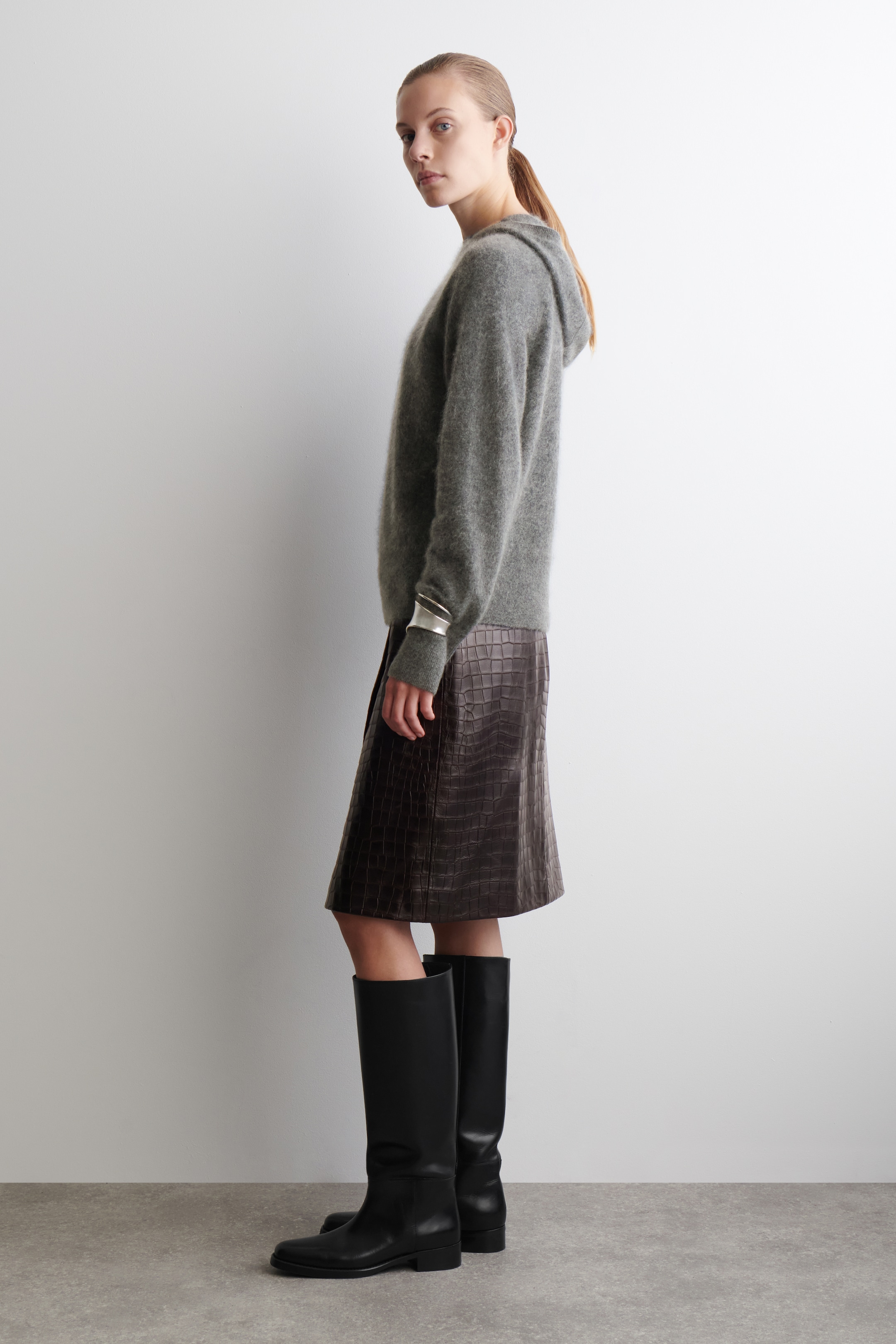 Ingrandisci l'immagine: Un maglione grigio con cappuccio, caratterizzato da una morbida texture in maglia spazzolata e una vestibilità rilassata, viene presentato di profilo. È abbinato a una gonna a trapezio in rilievo effetto coccodrillo di colore marrone scuro, stivali neri alti fino al ginocchio e un ampio bracciale a polsino color argento indossato sopra la manica sinistra del maglione.