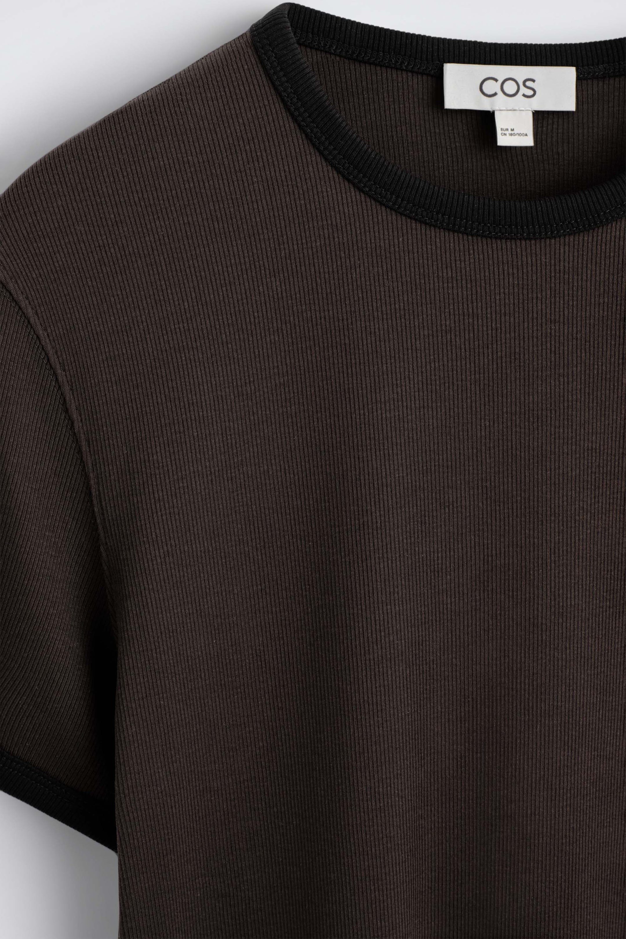 Ein schmales, geripptes Baumwoll-T-Shirt ist in einer Nahaufnahme von vorne zu sehen und zeigt einen dunkelbraunen Body mit feiner vertikaler Rippstruktur. Das Kleidungsstück hat einen kontrastierenden schwarzen, gerippten Rundhalsausschnitt sowie einen dazu passenden schwarzen Ärmelsaum. Ein schmales, geripptes Baumwoll-T-Shirt ist in einer Nahaufnahme von vorne zu sehen und zeigt einen dunkelbraunen Body mit feiner vertikaler Rippstruktur. Das Kleidungsstück hat einen kontrastierenden schwarzen, gerippten Rundhalsausschnitt sowie einen dazu passenden schwarzen Ärmelsaum.
