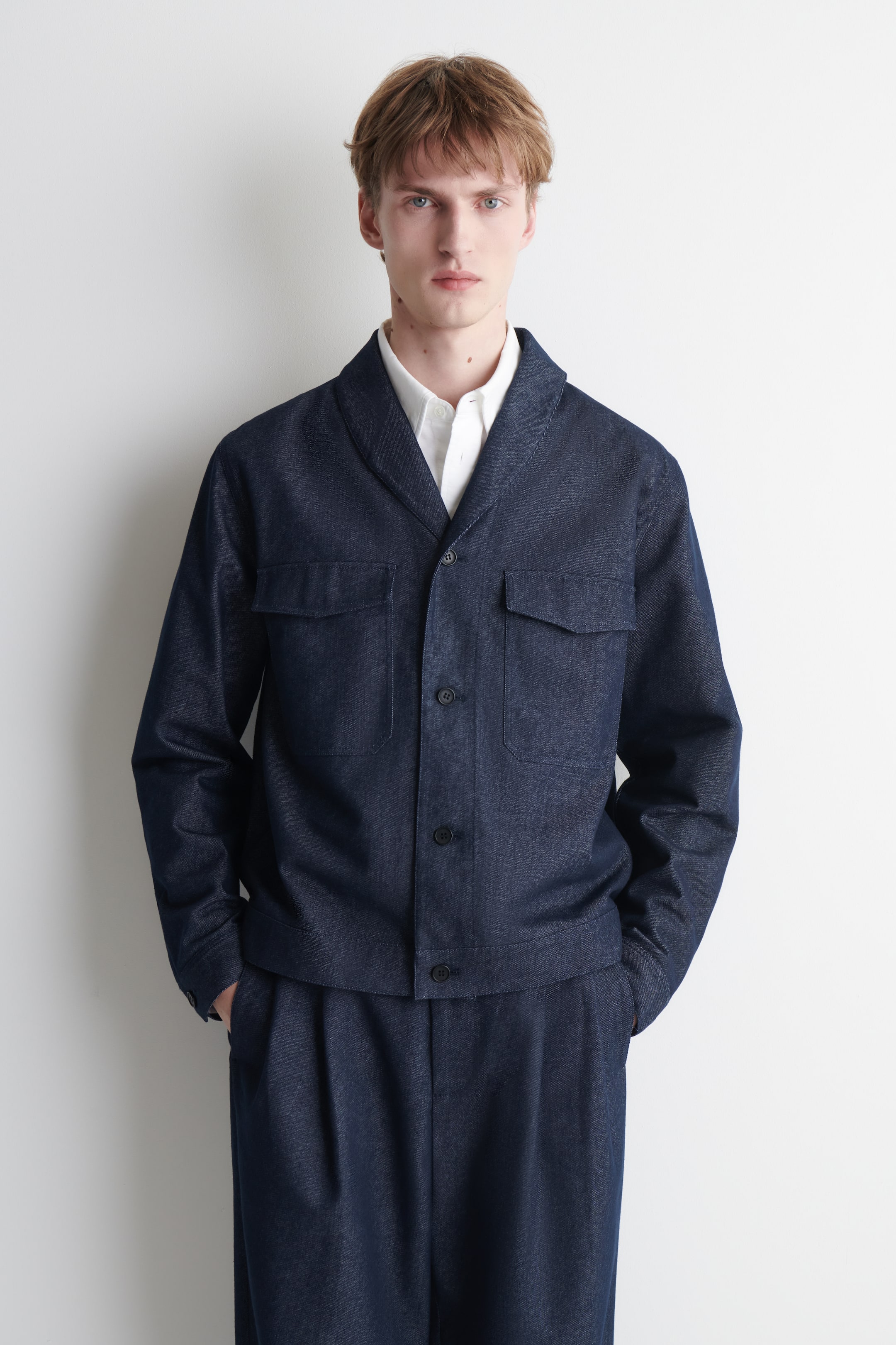 View larger image: A dark blue denim jacket is shown facing forward, featuring a shawl collar, a three-button front placket, and two flap chest patch pockets. It is layered over a white collared shirt and paired with matching dark blue pleated trousers.