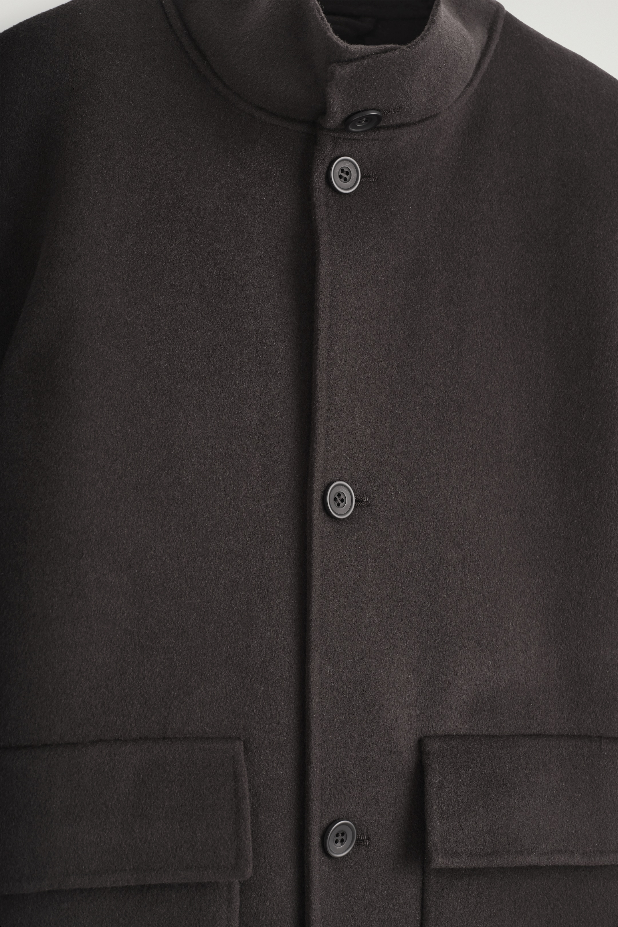 Grösseres Bild ansehen: Eine dunkelbraune Jacke wird von vorne präsentiert, mit einem hohen Trichterkragen, einem zentralen Knopfverschluss mit dunklen Knöpfen und zwei großen Klapptaschen auf der Vorderseite.