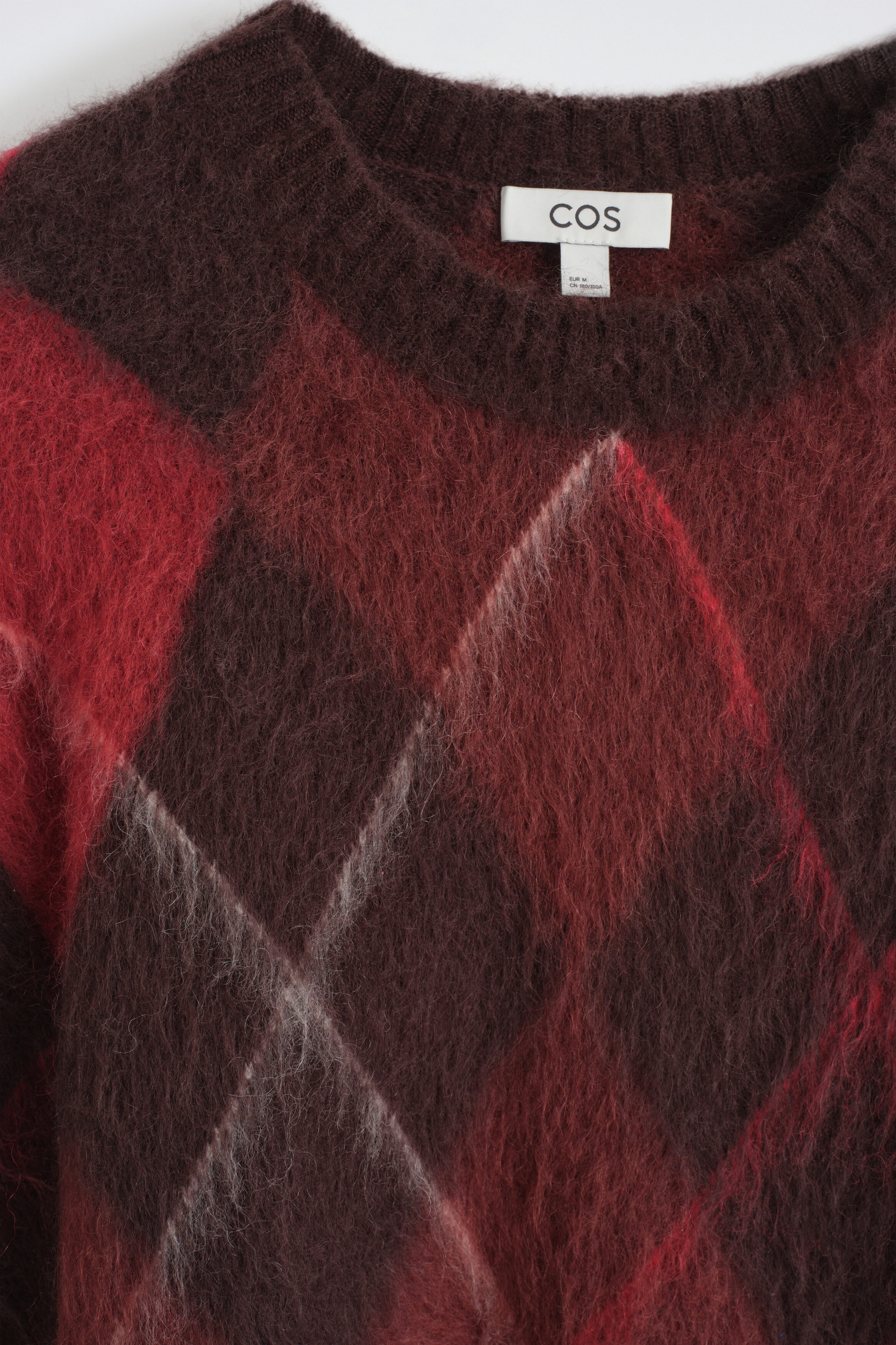 Ein flauschiger Strickpullover präsentiert ein markantes Argyle-Muster in Dunkelbraun und Rot mit feinen, sich kreuzenden hellgrauen Linien. Die Nahaufnahme zeigt einen gerippten Rundhalsausschnitt und einen weichen, strukturierten Stoff mit einem weissen Markenetikett, das am inneren Halsausschnitt sichtbar ist.