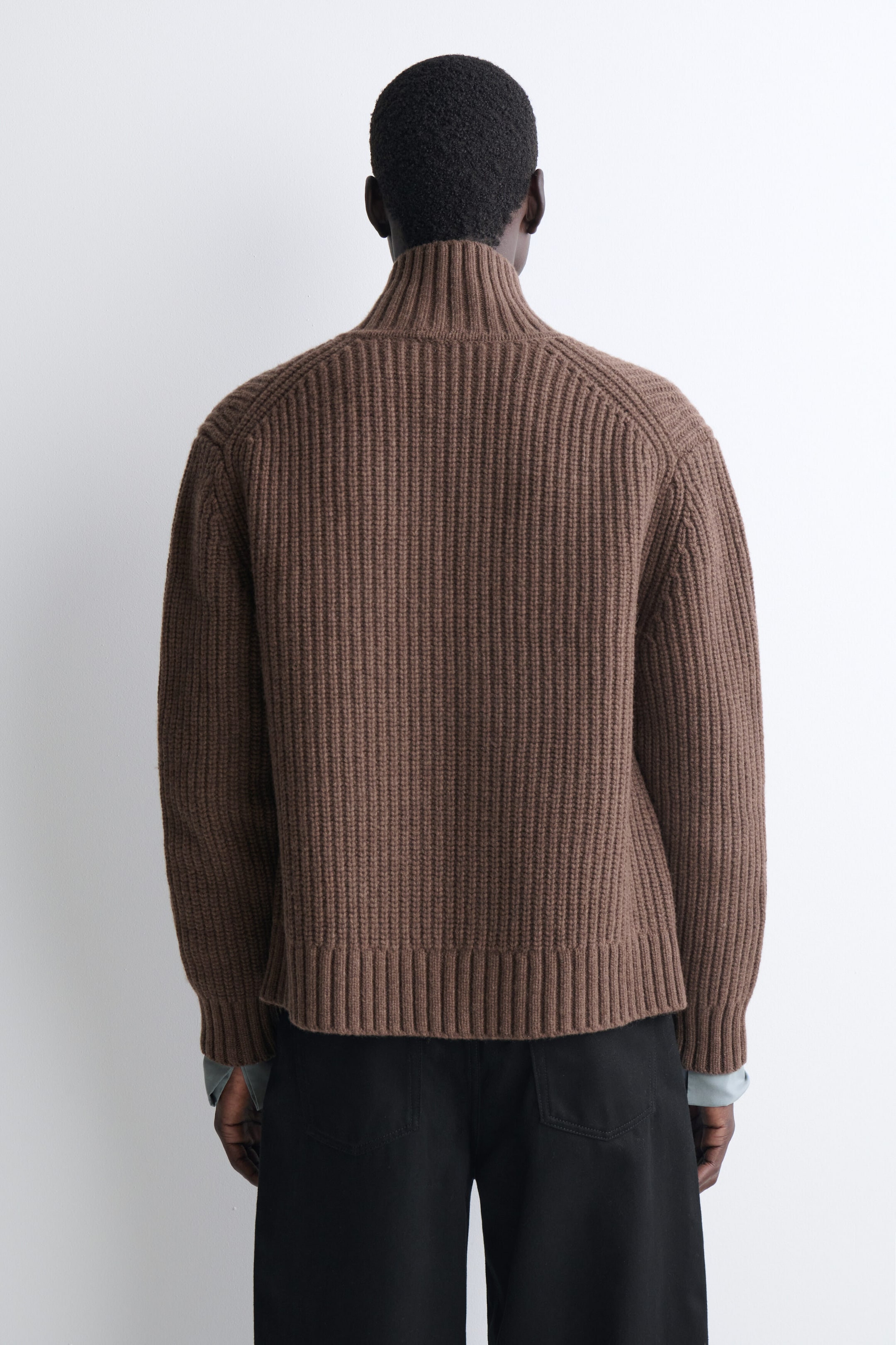 A brown ribbed-knit cardigan is shown from the back, featuring a high funnel neck, raglan sleeves, and matching ribbed hem and cuffs. The cardigan has a relaxed fit and is layered over black trousers. A brown ribbed-knit cardigan is shown from the back, featuring a high funnel neck, raglan sleeves, and matching ribbed hem and cuffs. The cardigan has a relaxed fit and is layered over black trousers.