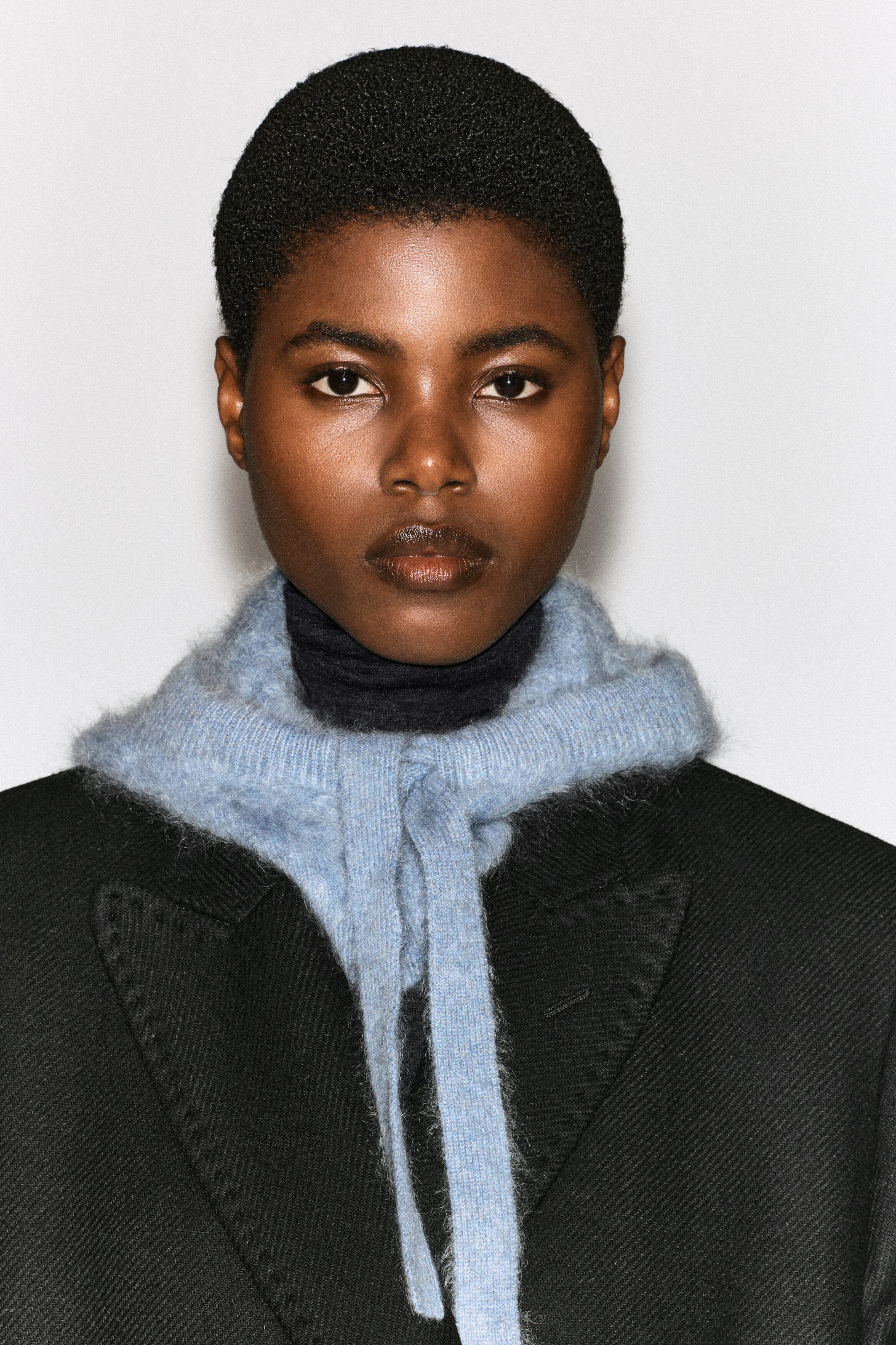 A light blue brushed cashmere hood is worn as a collar around the model’s neck, layered over a dark turtleneck and a black textured coat. The hood features a soft, textured finish and long matching knitted ties that hang down the front.