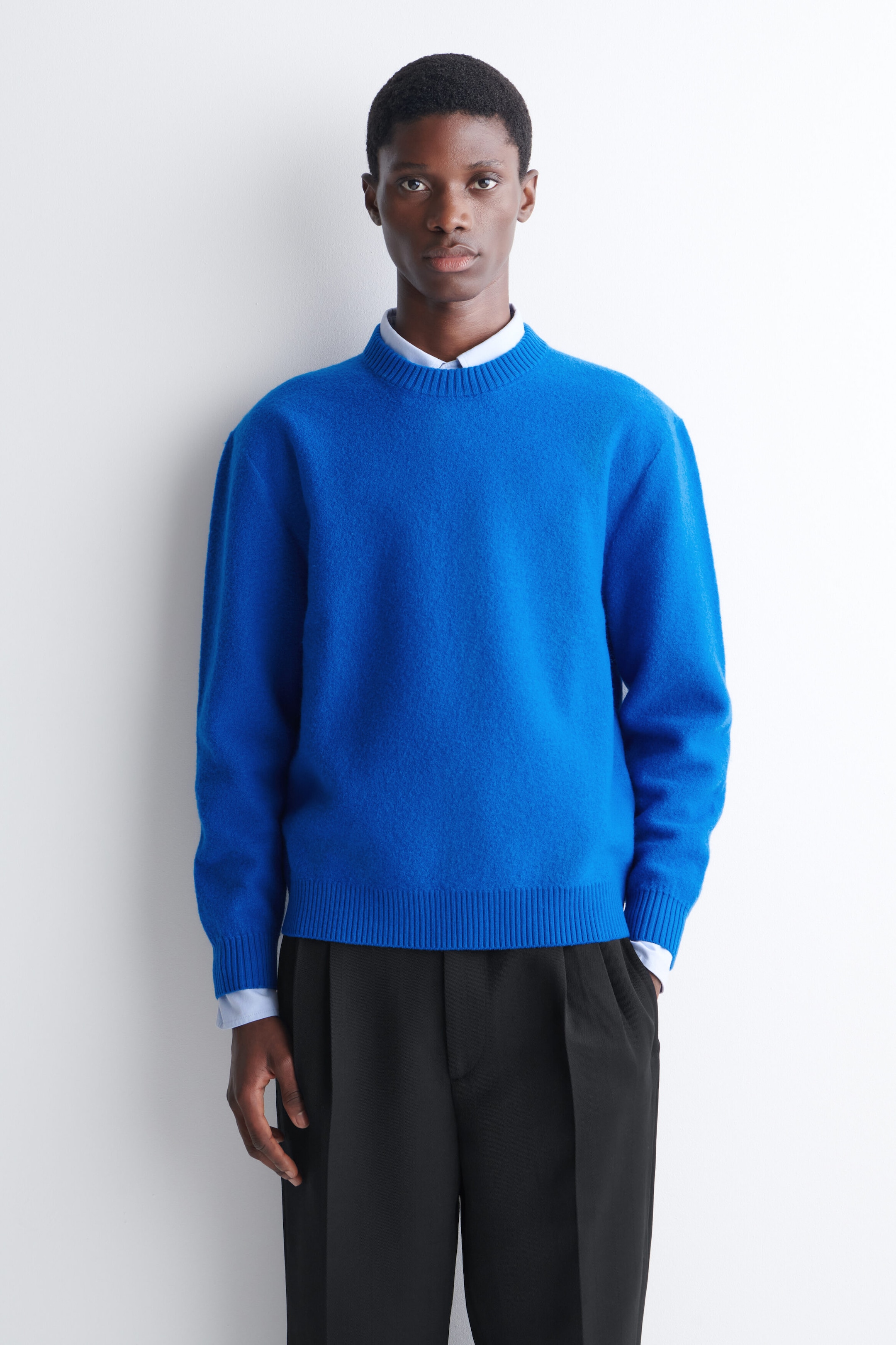 A bright blue boiled wool sweater is worn by the model, layered over a light blue collared shirt and paired with black trousers. The sweater features a crew neck and ribbed trims at the collar, cuffs, and hem. A bright blue boiled wool sweater is worn by the model, layered over a light blue collared shirt and paired with black trousers. The sweater features a crew neck and ribbed trims at the collar, cuffs, and hem.