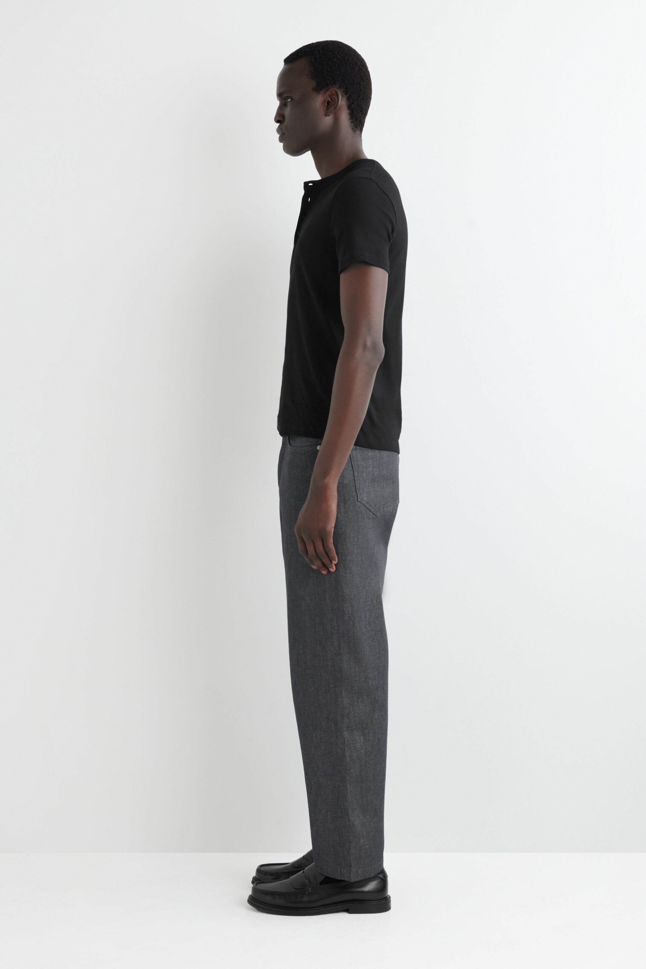 View larger image: A black short-sleeve Henley shirt with a button placket at the neckline is being shown in a side profile, paired with dark gray straight-leg pants and black leather loafers with a thick sole.